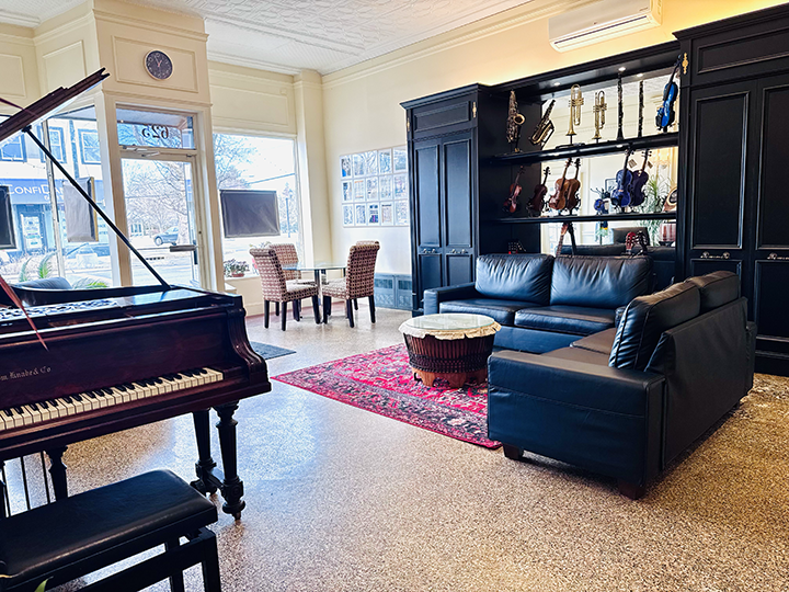 Living room with black leather sofa, grand piano, red area rug, acoustic guitars and violins displayed on black shelving, and a small dining area with two chairs near windows.