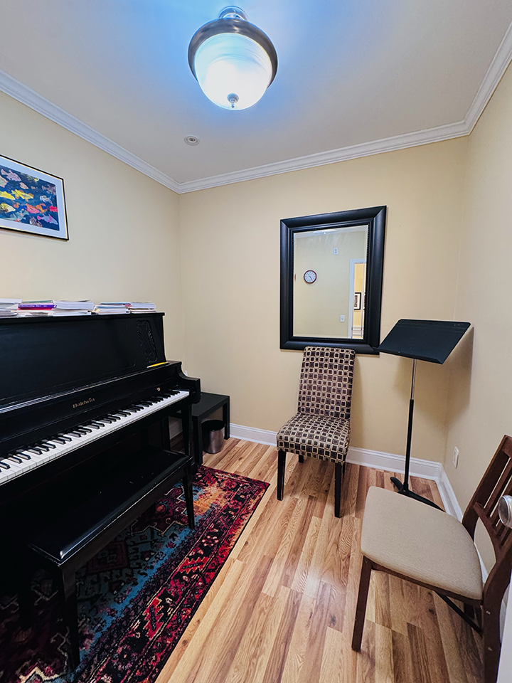 A small music room with a black upright piano, a colorful patterned rug, three chairs, a wall mirror, a framed picture of fish, and a music stand. The walls are beige, and the room has hardwood floors.