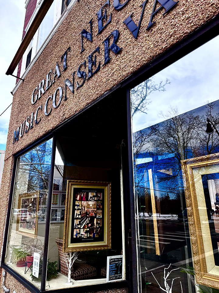 Exterior of Great Necks Music Conseravtory with large window displays featuring framed artwork and indoor plants, reflecting trees and street scene outside.