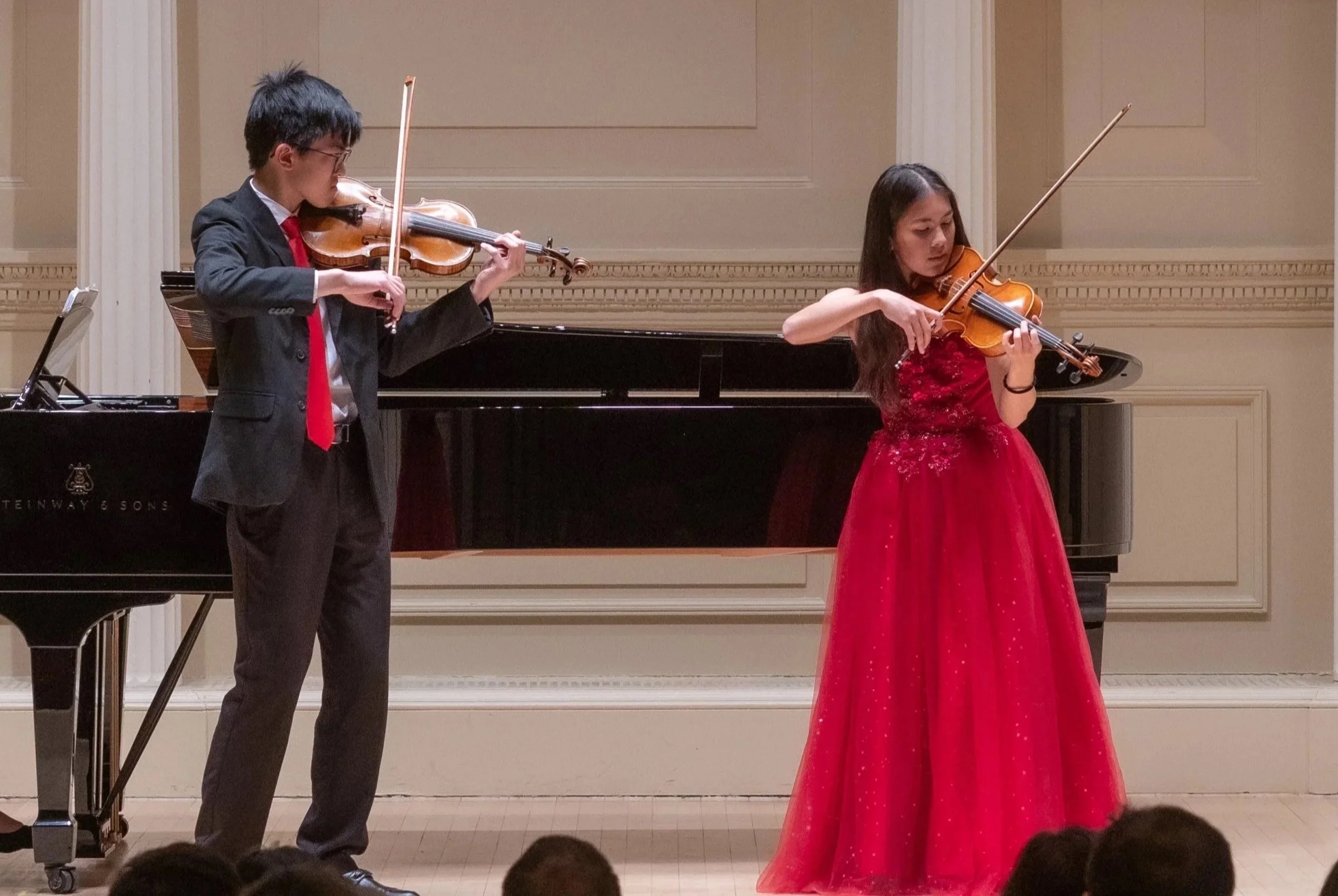 Two young musicians performing with violins on stage, one male and one female, with the female in a red dress and the male in a black suit with a red tie, in front of an audience.