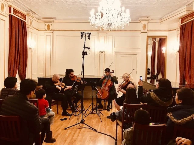 A string quartet performing in an elegant room with an audience watching, some recording on phones, and a chandelier hanging above.