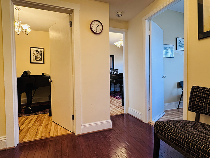 A hallway with three doorways, one leading to a room with a piano, another to a room with a desk and framed artwork, and a third to a room with blue walls and a chair. A clock is on the wall, and there is a checkered chair on the right.