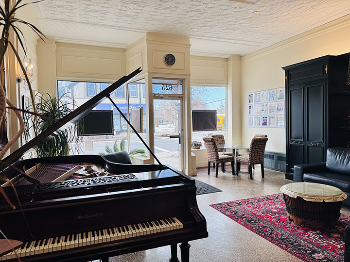 Interior of a cozy living room with a black grand piano, a large window with two television screens, a black cabinet, a small round coffee table, a red patterned rug, and two chairs near the window.