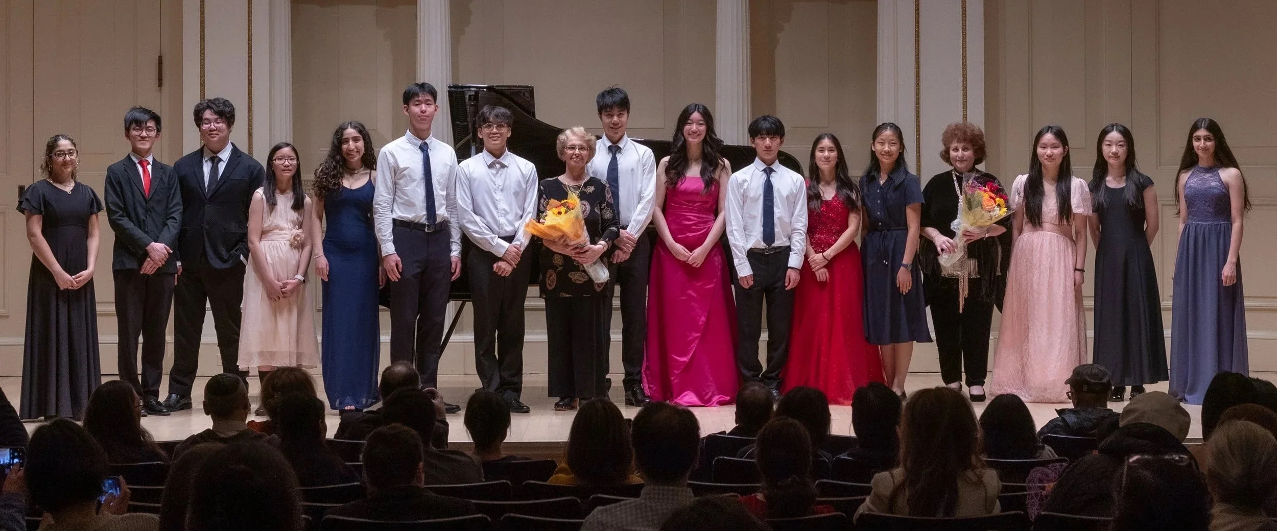 Group of people on stage after a performance, with some holding bouquets of flowers, in a concert hall with an audience in front.