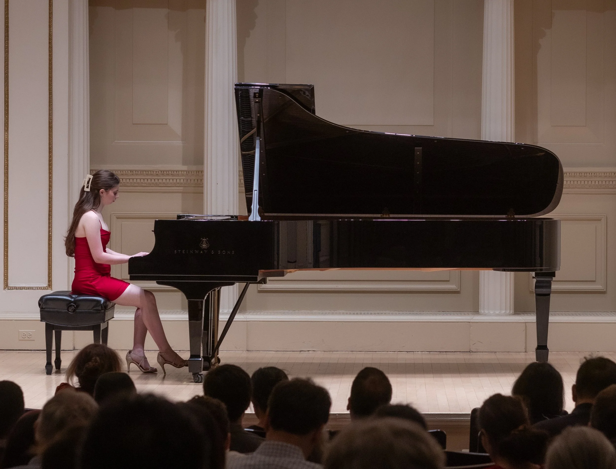 A young woman in a red dress playing a black grand piano on a stage in front of an audience.