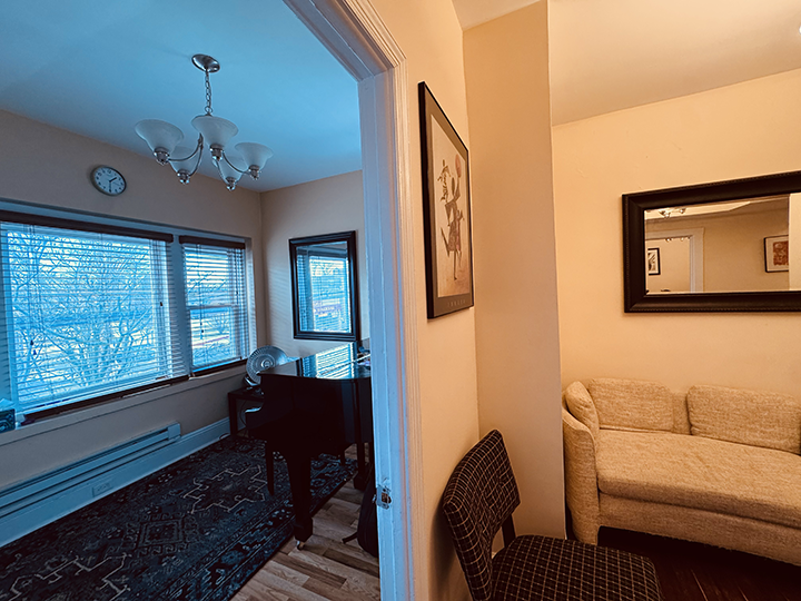 A view of two rooms in a house, separated by a doorway. The room on the left has large windows, a corner piano, a chandelier, and a clock on the wall. The room on the right has a beige sofa, a large mirror on the wall, and a framed picture. There is 