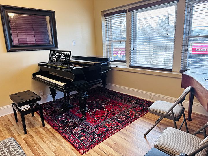 Room with a black upright piano on a red patterned area rug, a small black bench in front, and gold chairs nearby. There are three windows with blinds, a large mirror on the wall, and a wooden floor.