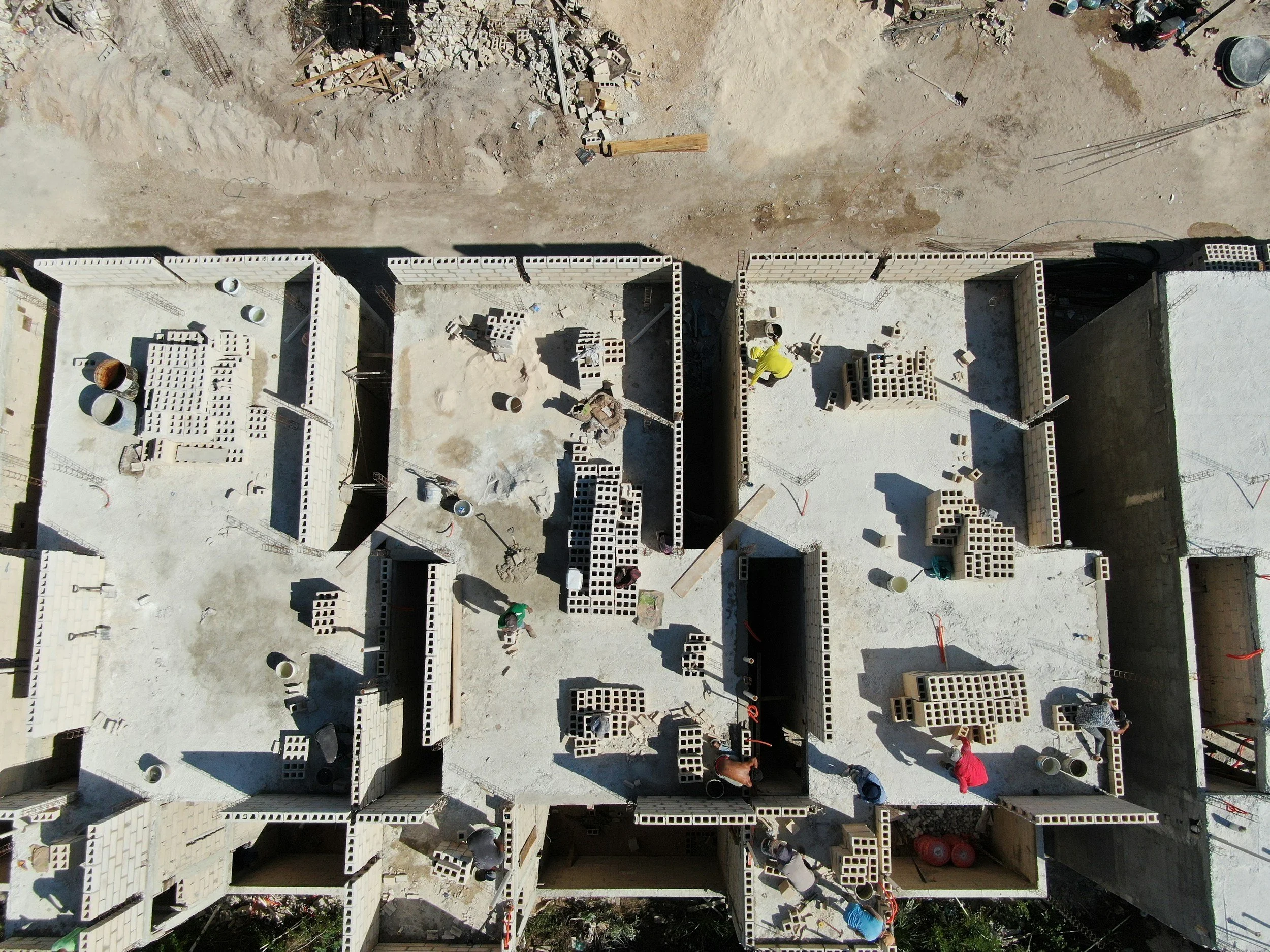 An aerial view of a construction site showing several partially built brick structures with workers actively working inside and around them.