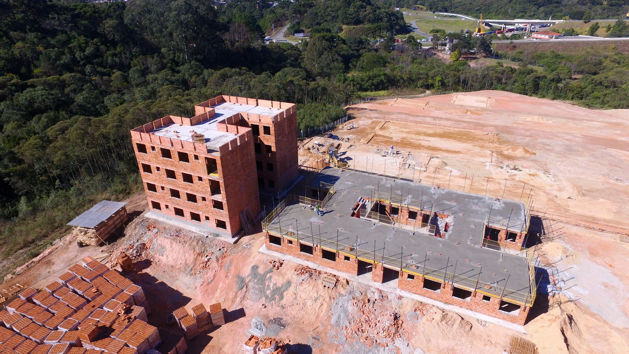 Aerial view of a construction site with partially built brick buildings, surrounded by open land and trees, with a highway and forest in the background.