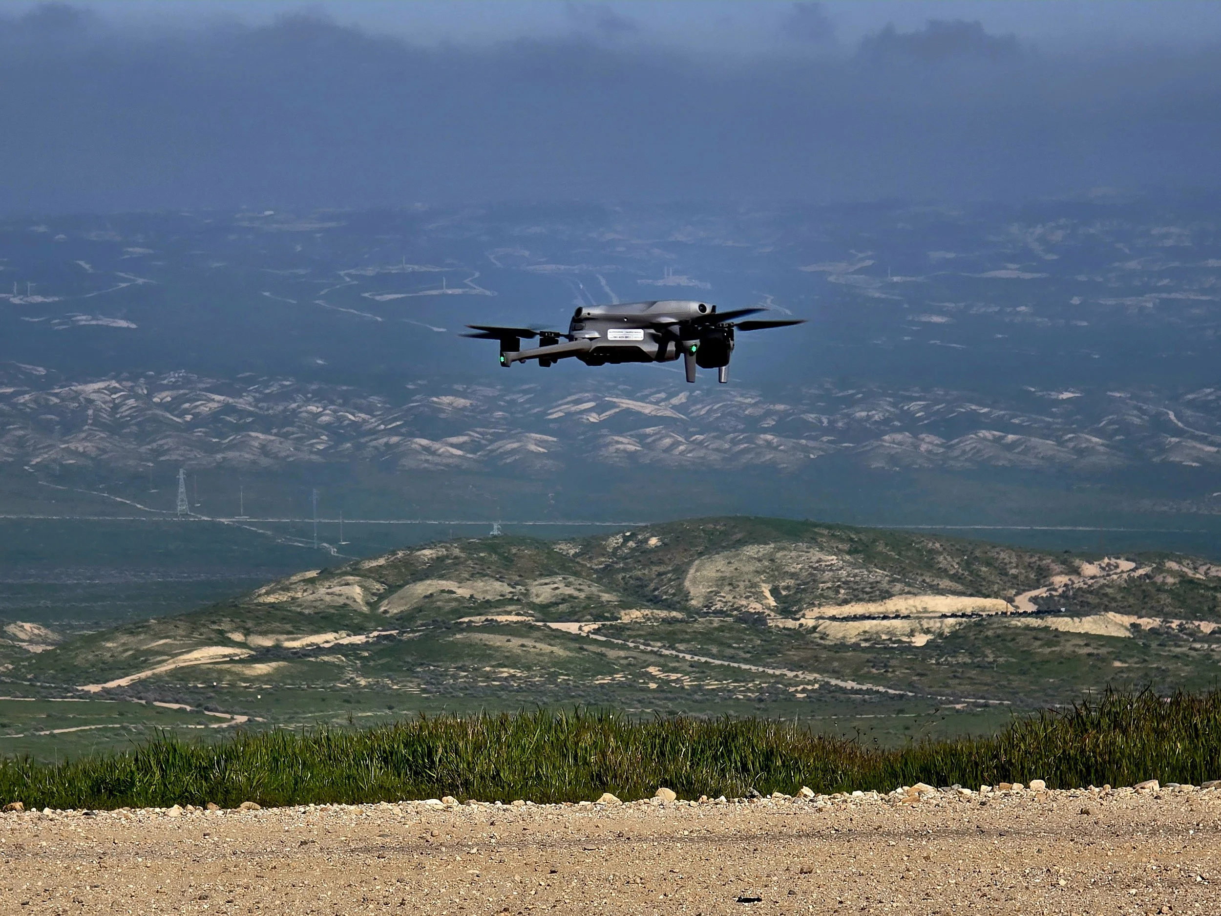 A drone flying over grassy and hilly terrain with mountains in the background under a cloudy sky.
