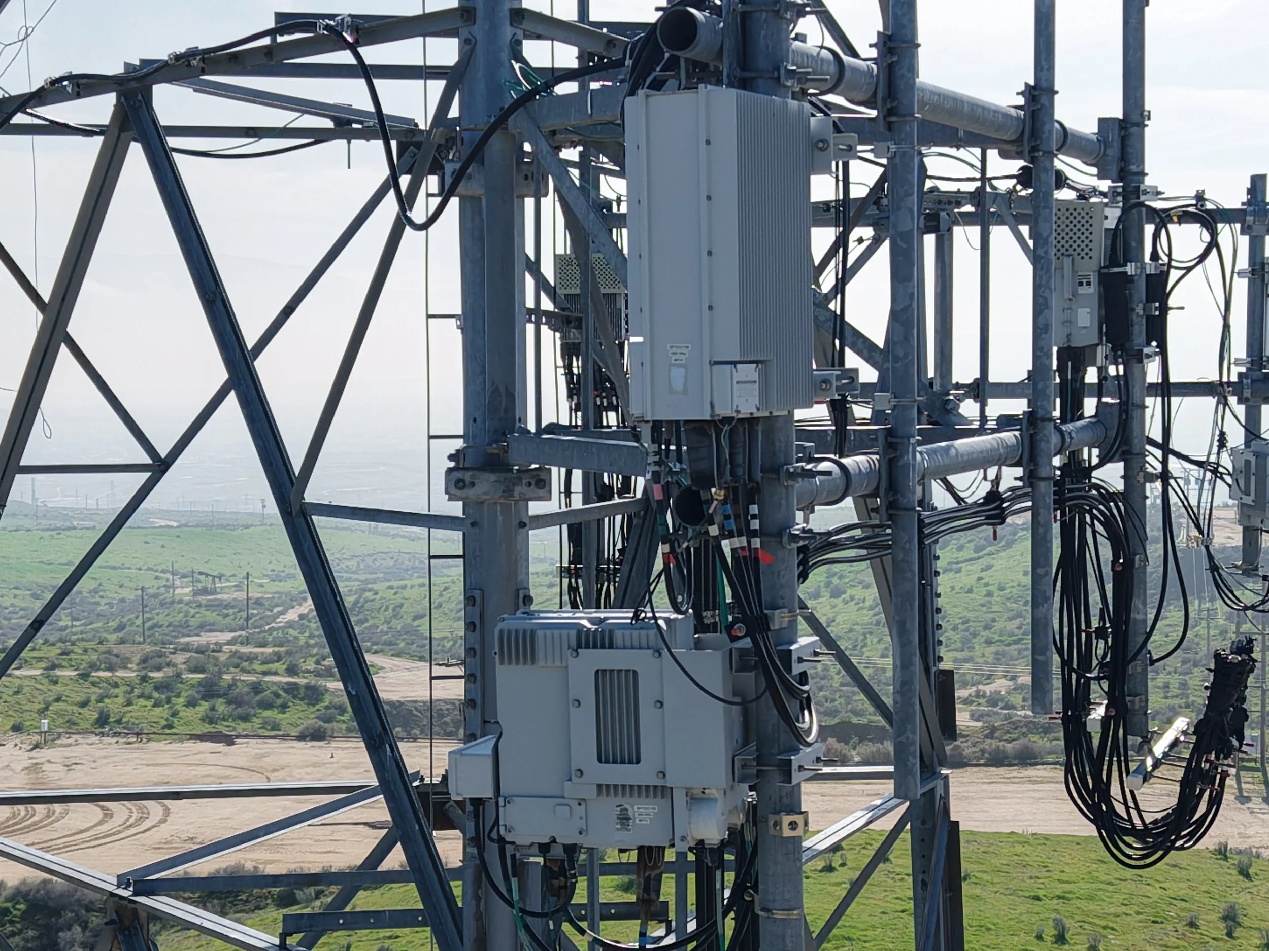 Close-up of a telecommunications tower with electrical and signal equipment, wires, and pipes, set against a landscape of fields and hills.