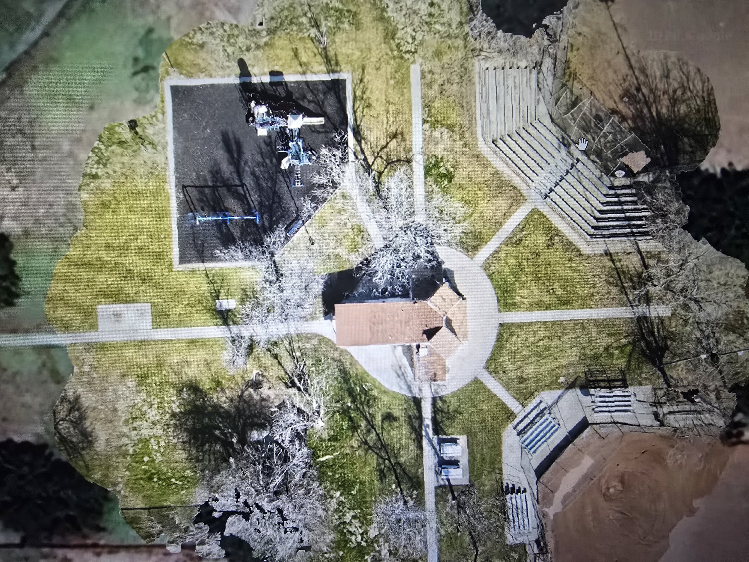 An aerial view of a park with a circular paved area, a small building, playground equipment, and tiered amphitheater seating, with pathways connecting these features.