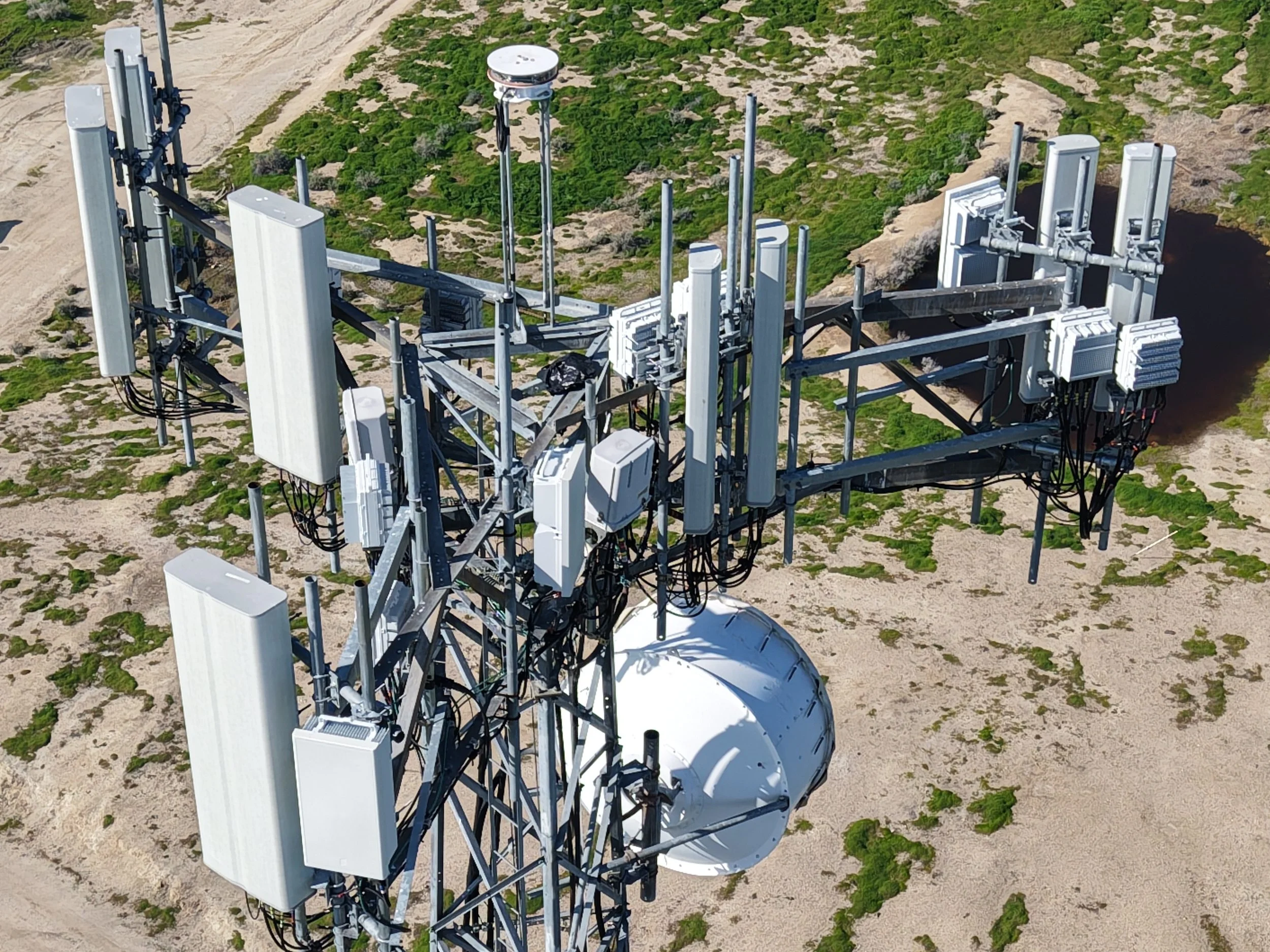 Aerial view of a telecommunications tower with multiple antennas and equipment on a sandy and grassy landscape.