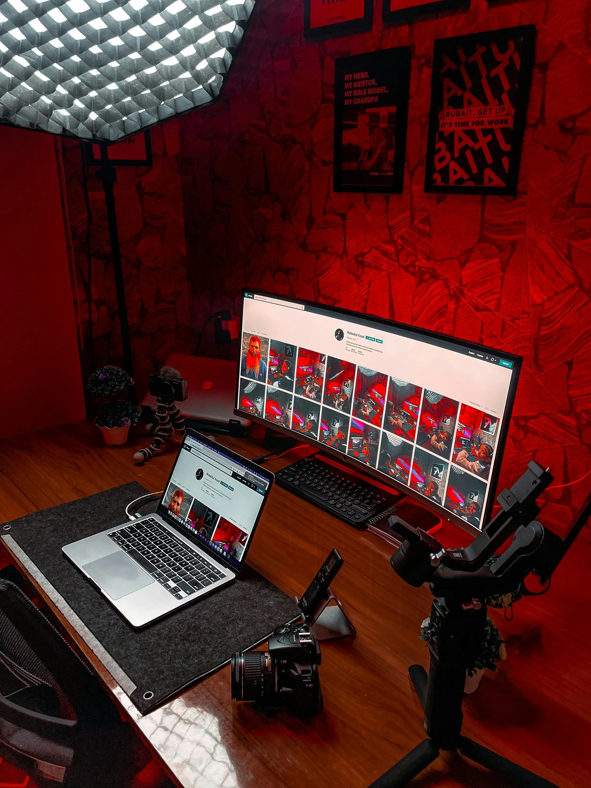 Immersive dark-room setup lit by red LEDs. Large ultawide monitor shows grid of video thumbnails featuring a red-haired man. Open MacBook displays creator profile. Surrounded by soft boxs, gimbal camera, mic, and motivational posters.