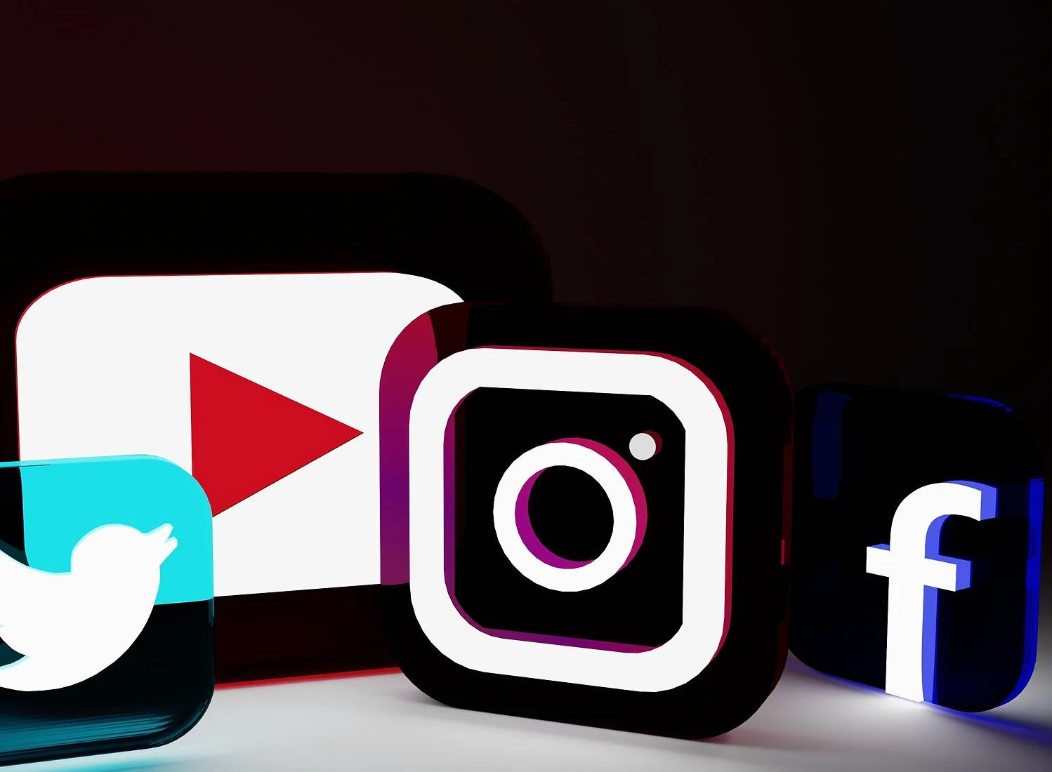 Dramatic Studio shot of glowing #D social media logos on black background: blue twitter bird, red Youtube play button, purple-pink Instagram camera, and blue Facebook 'f". Each Icon casts Colourful neon flows and shadows.