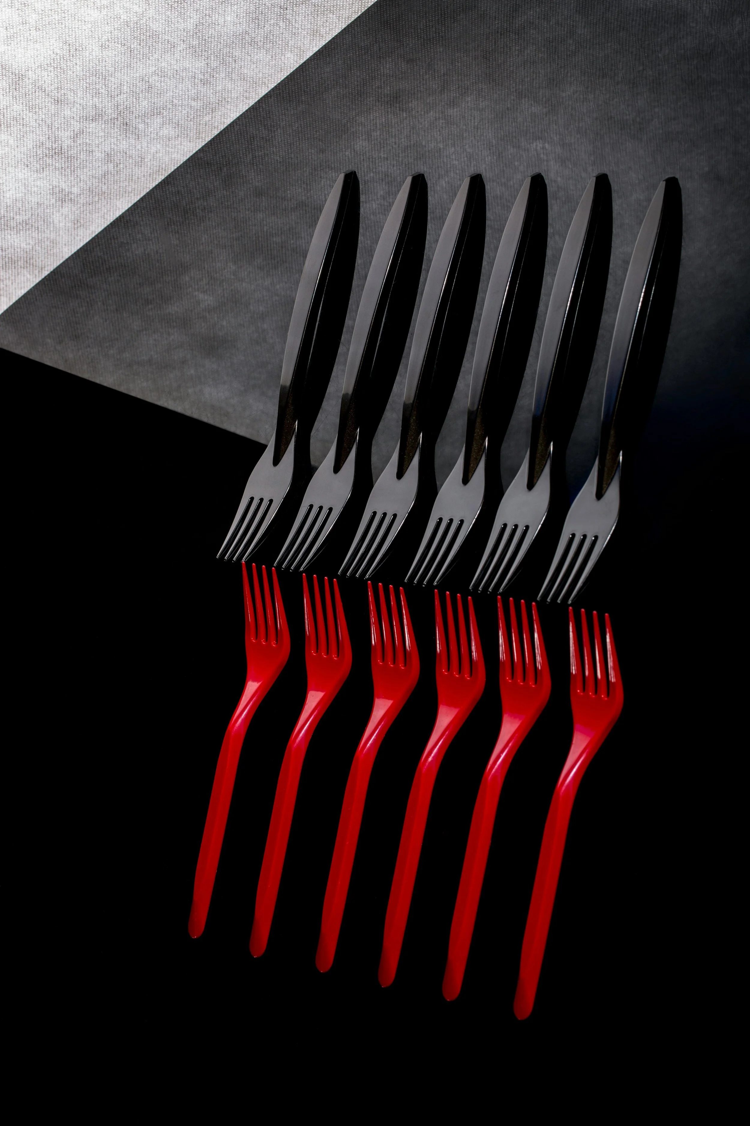 Ten Sleek modern forks in two rows on dark textured surface. Five Matte black forks and five glossy red forks create mirror-like reflections underneath. High Contrast side lighting against gradient gray-black background.