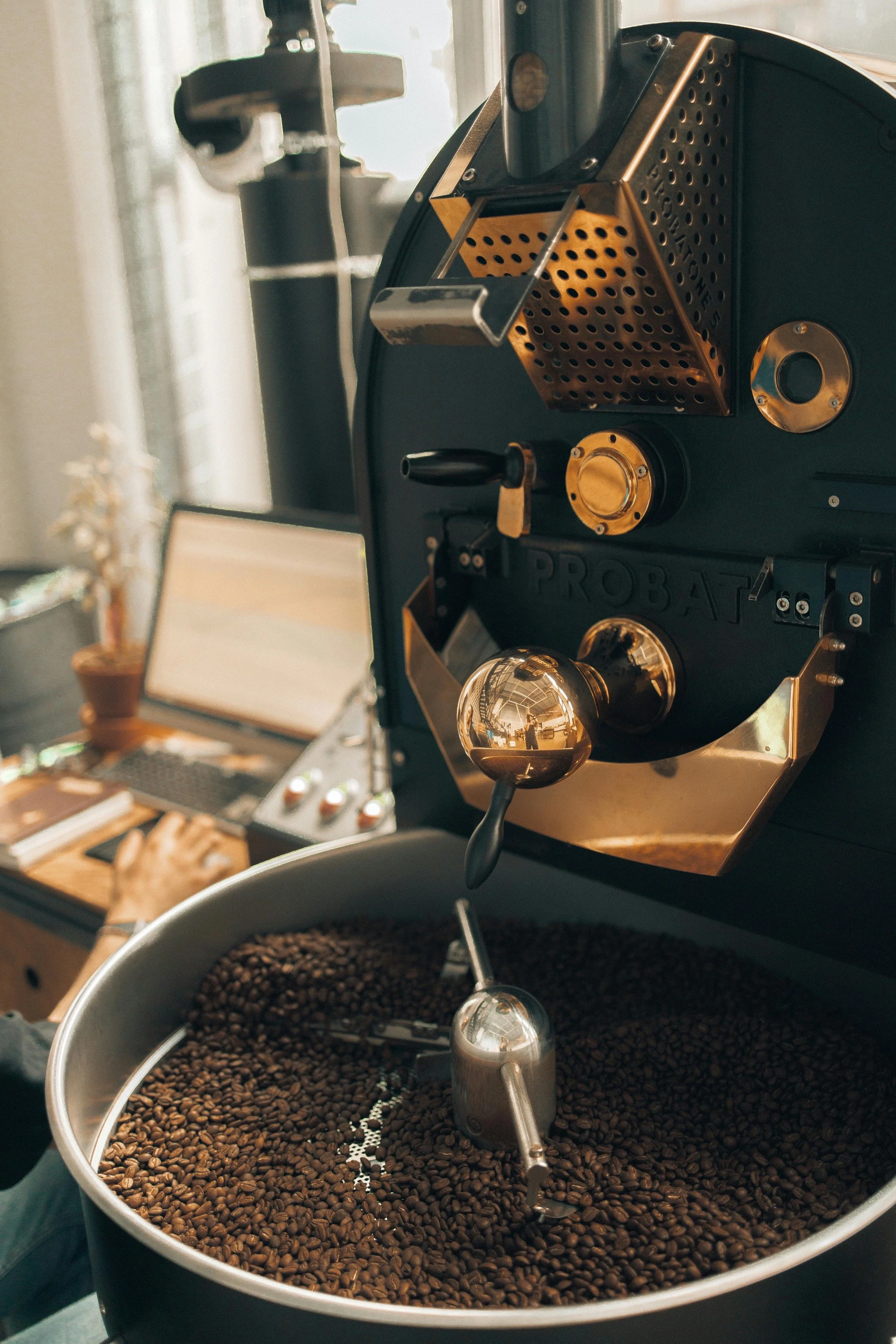 Close-up of a coffee roasting machine with roasted coffee beans in a large metal drum, while a person works on a laptop in the background.