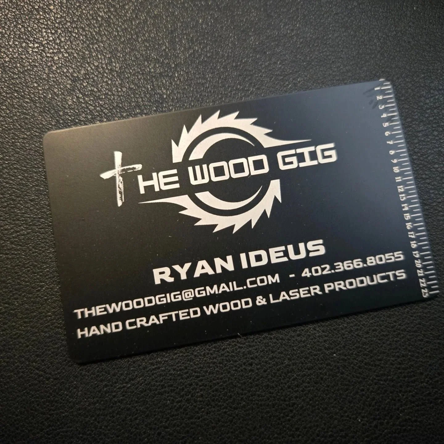 Black business card for The Wood Gig with white text displaying contact information, email, phone number, and products, placed on black textured surface.