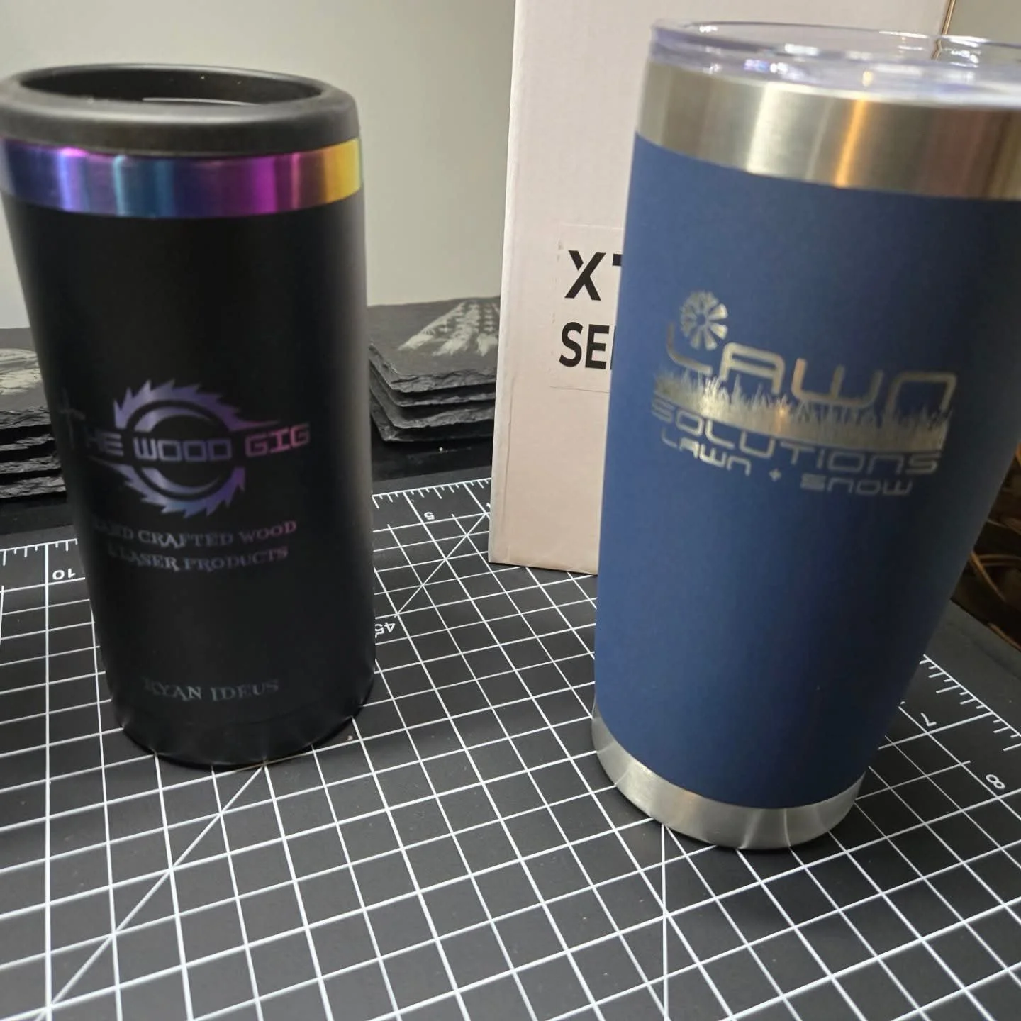 Two travel mugs with logos on a cutting mat with a grid, a white box behind them, and a stack of black material in the background.