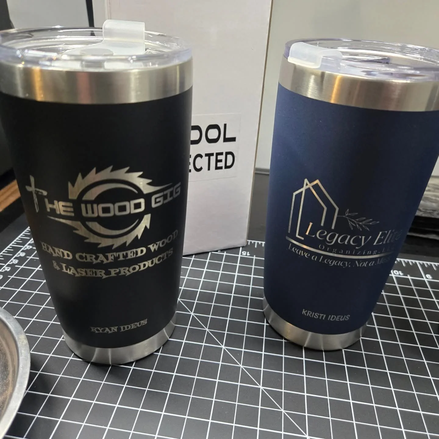 Two stainless steel tumblers with printed logos, one black with 'The Wood Gig' logo and the other blue with 'Legacy Elite Organizing' logo, placed on a black grid cutting mat.