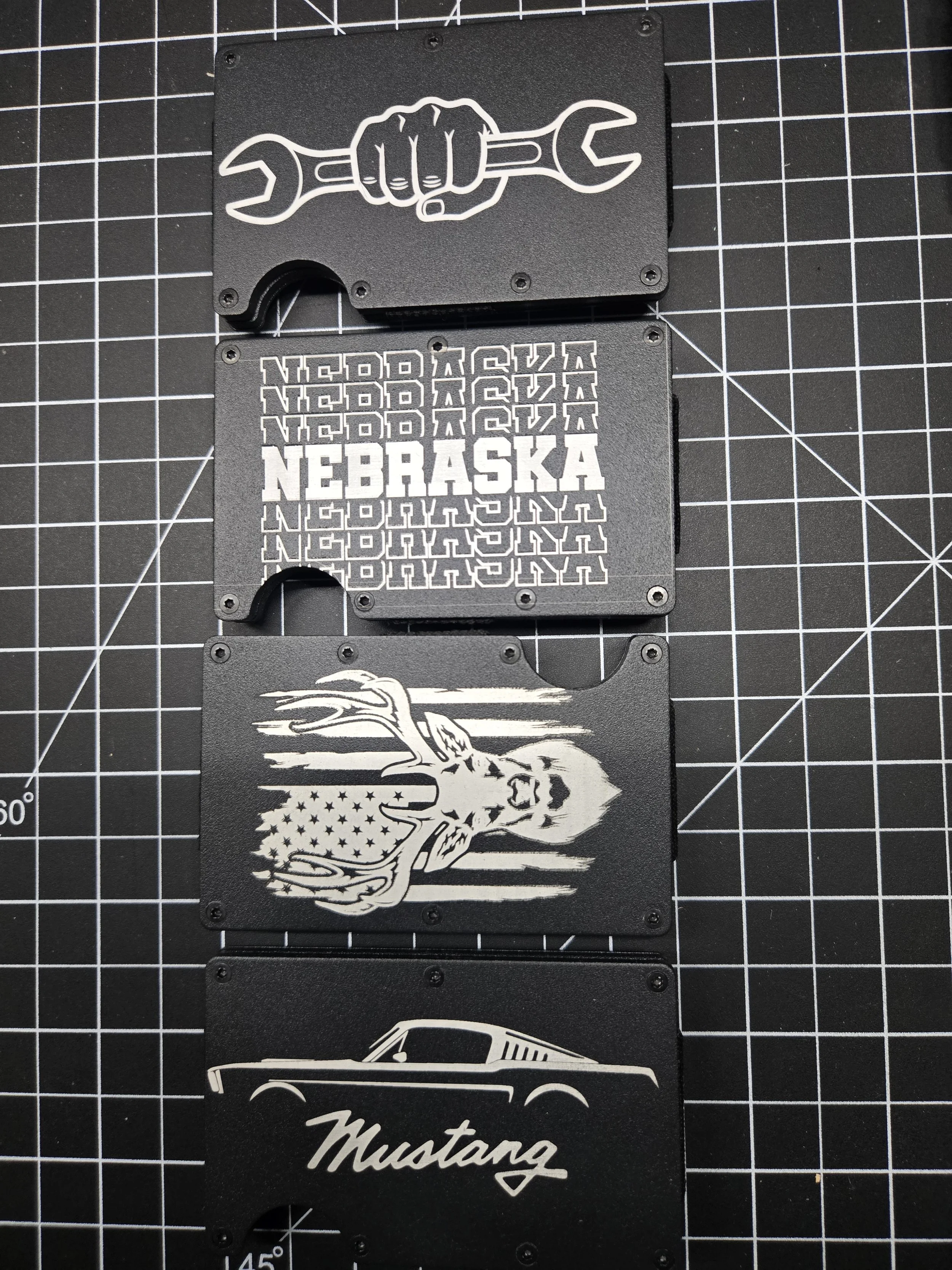 Four black metal cards with white designs and text are laid out on a cutting mat. The top card features a hand gripping a wrench. The second card displays the word Nebraska repeated multiple times, with one instance in bold across the middle. The thi