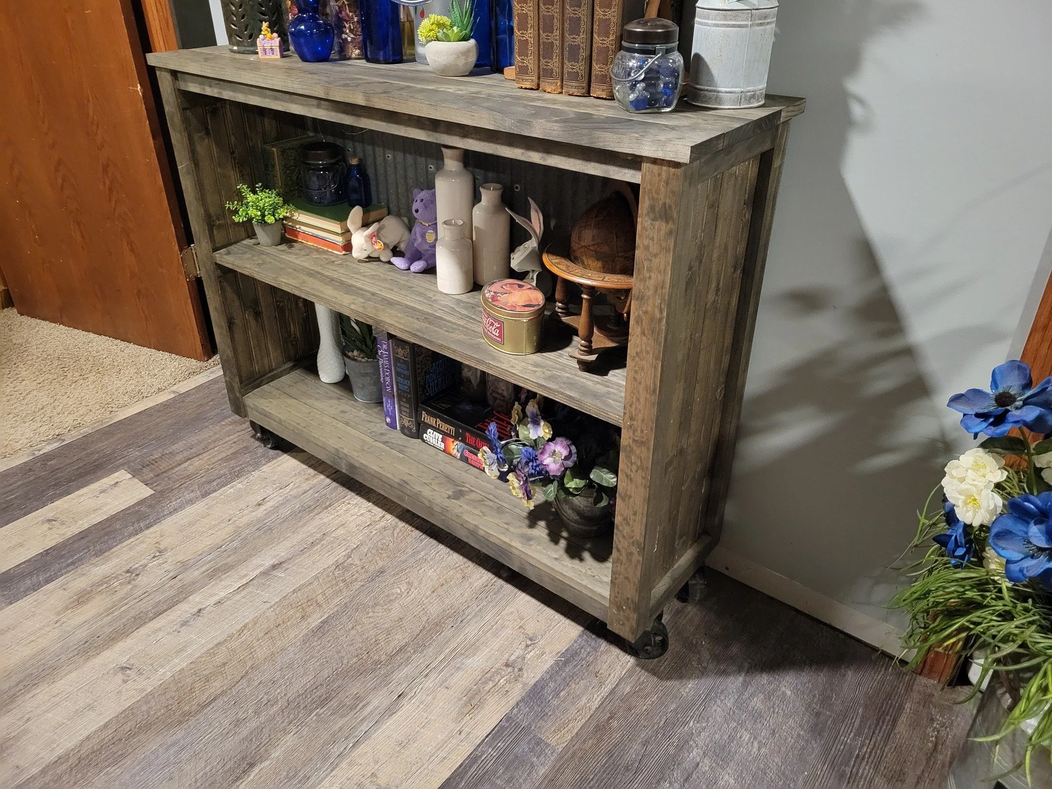 Wooden bookshelf on wheels with decorative items, books, and plants, against a wall with hardwood and tile flooring.