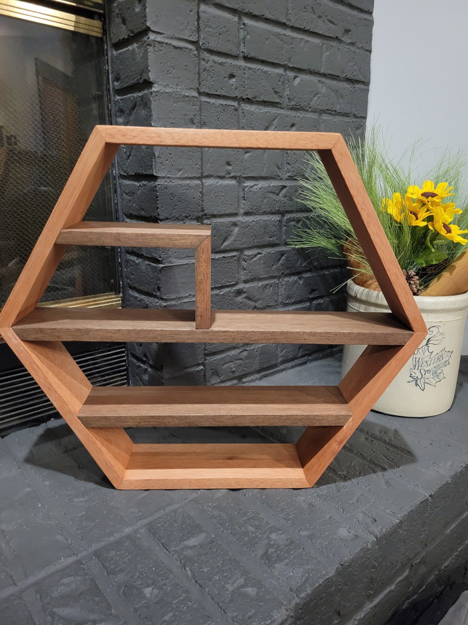 Wooden geometric bookshelf with hexagonal frame, placed near a brick fireplace and a potted plant with yellow flowers.