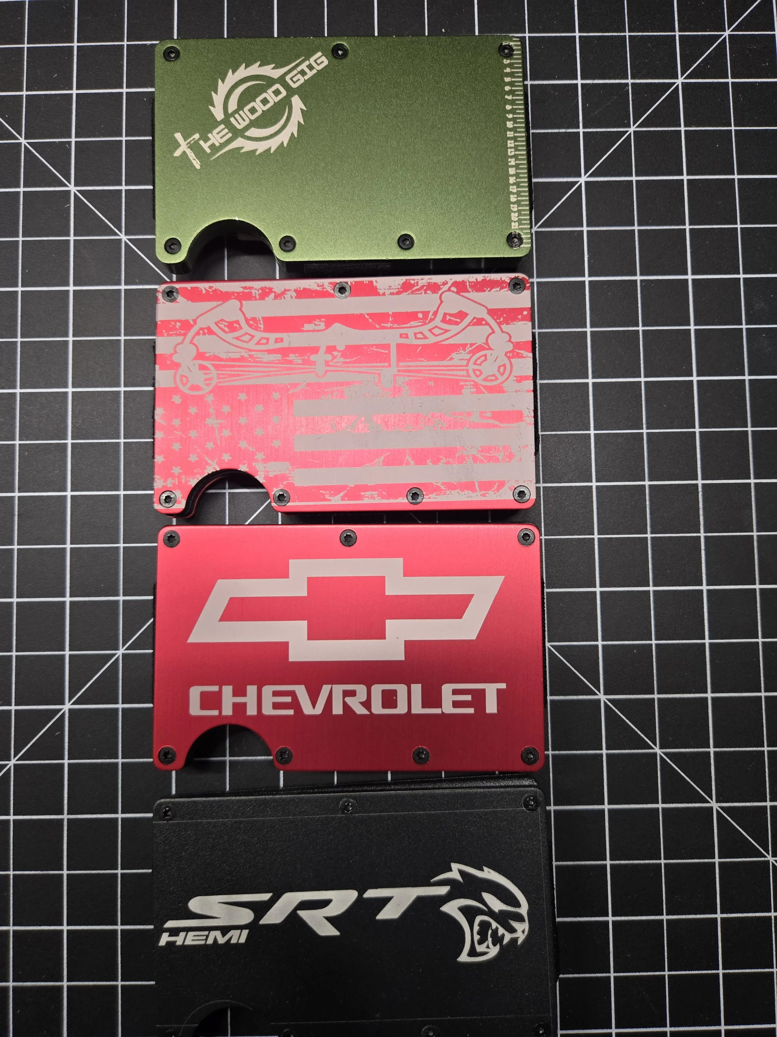 Four metallic card covers with automotive brand logos and graphics, arranged vertically on a measuring grid. The covers feature logos for The Wood GTG, Chevrolet, and SRT HEMI.