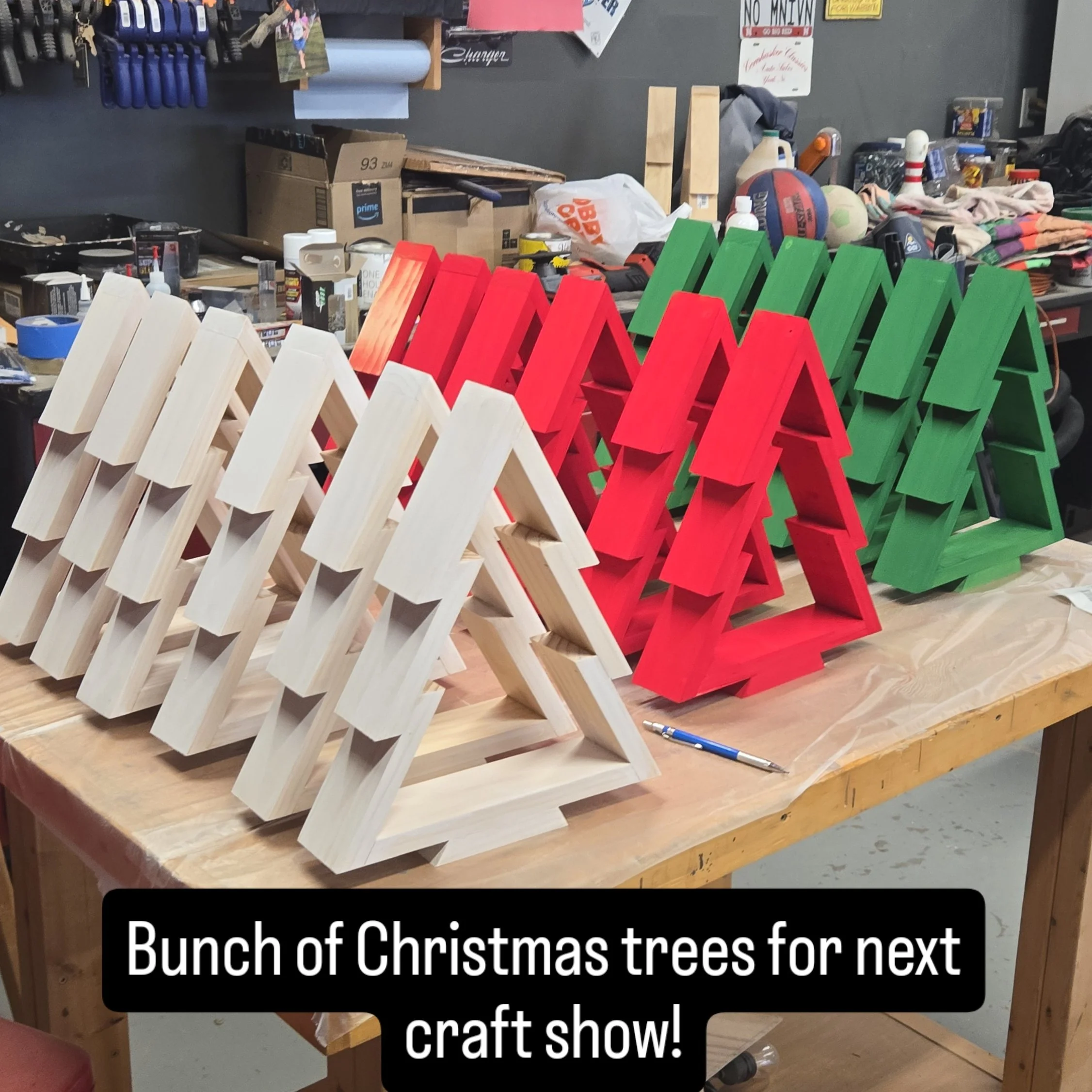 Colorful wooden Christmas tree decorations in natural, red, and green, arranged on a workbench for a craft show, with crafting supplies and tools in the background.