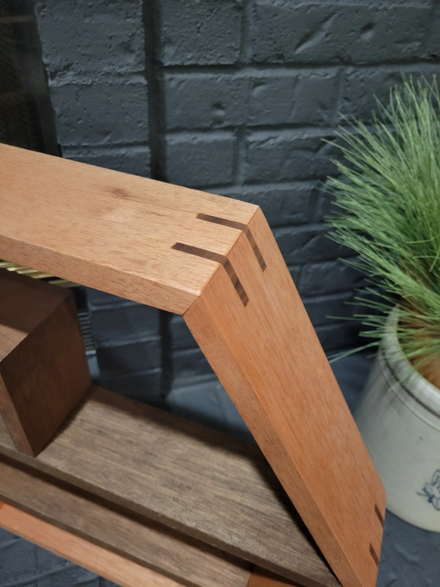 Close-up of a wooden furniture corner with dovetail joints visible, placed on a wooden surface next to a potted plant with green grass-like leaves, against a dark gray brick wall background.