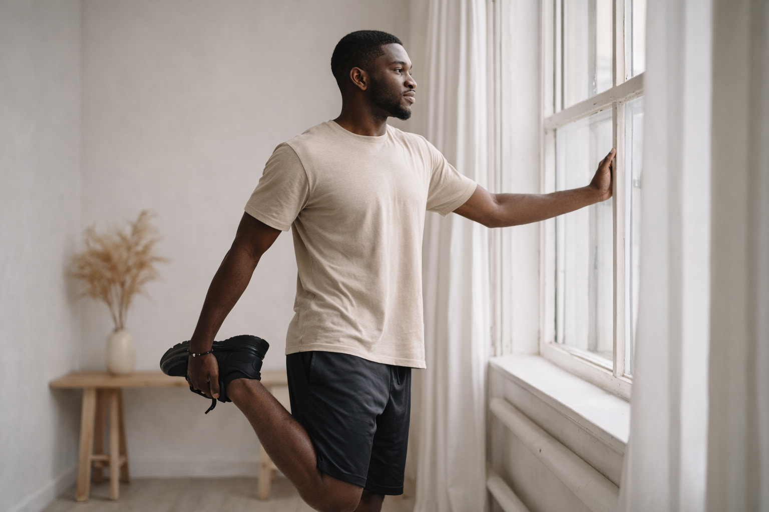 Man stretching by a window in a bright room, holding his foot behind his leg.