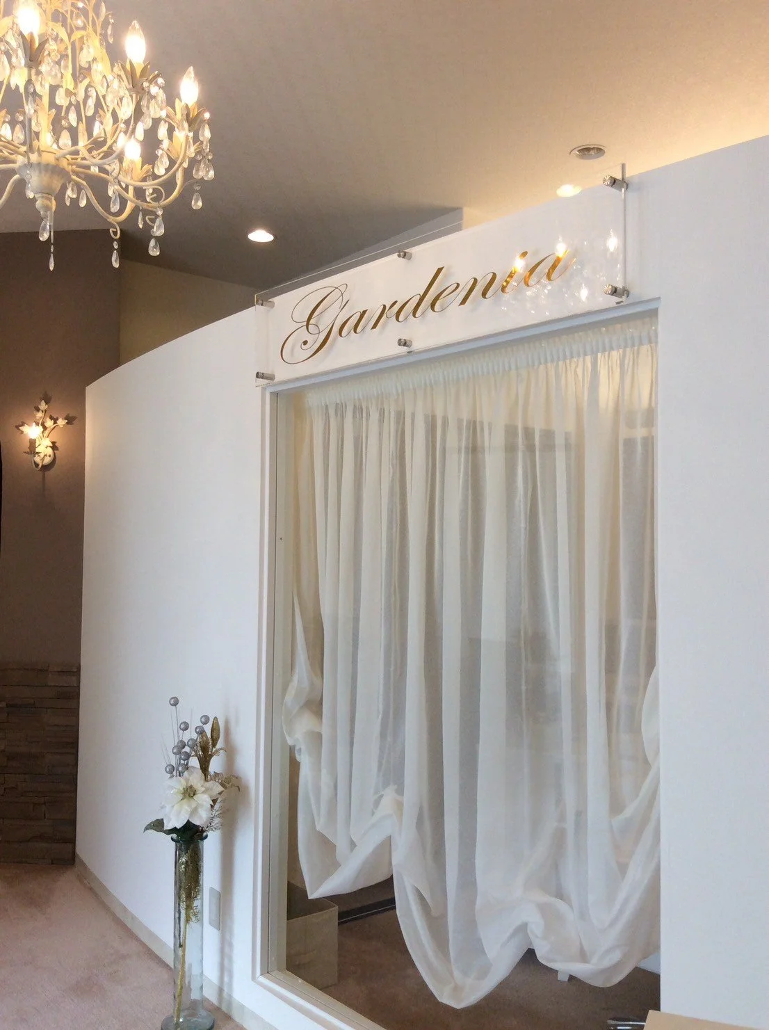 白いカーテンと花瓶に入った花が飾られた部屋の入り口。壁には「Gardenia」というサインがあり、上部にクリスタルが飾られたシャンデリアがついている。