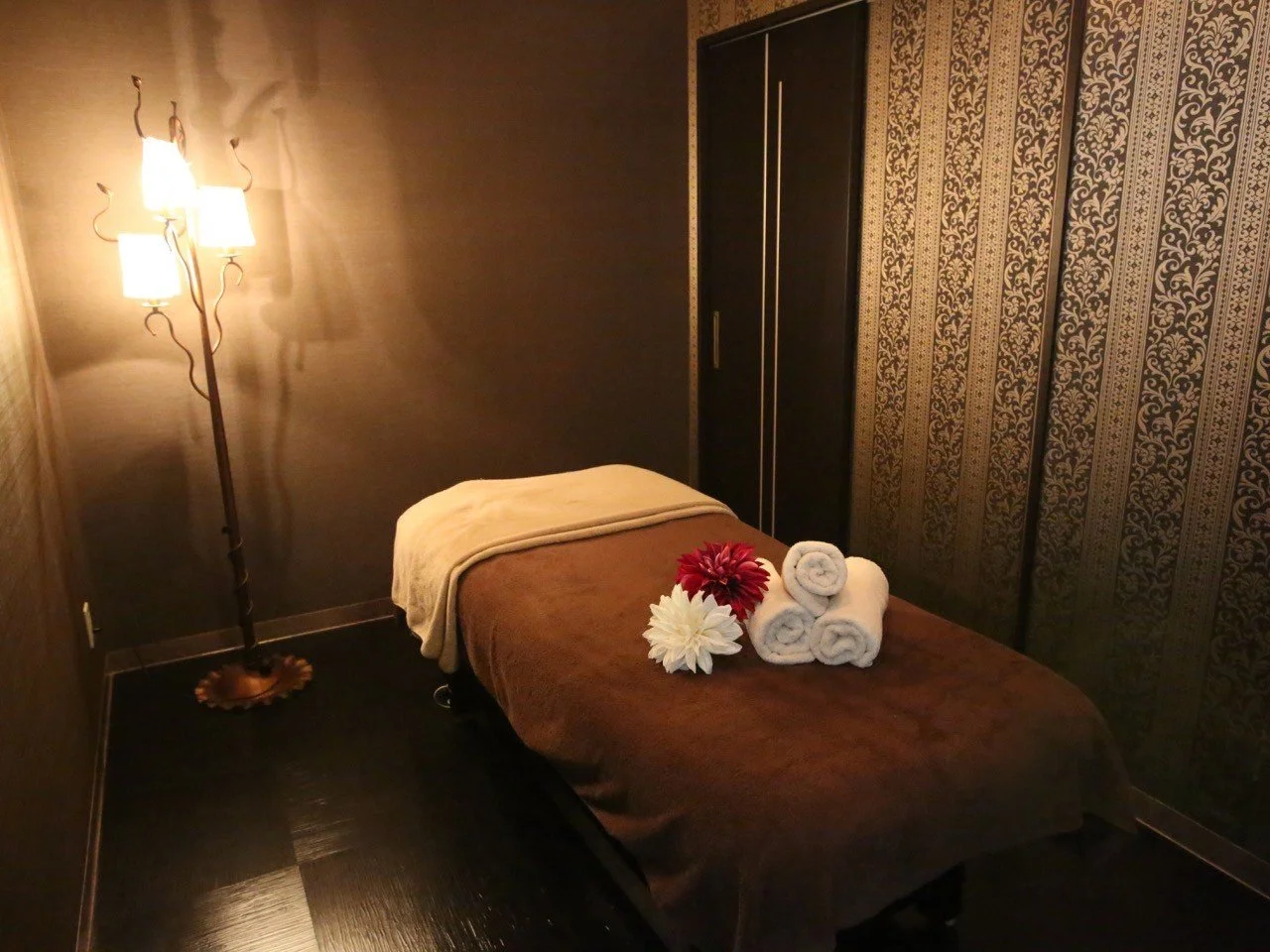 照明のついたリラクゼーションルーム、中央にマッサージベッド、上に折りたたみタオルと花が置かれている。