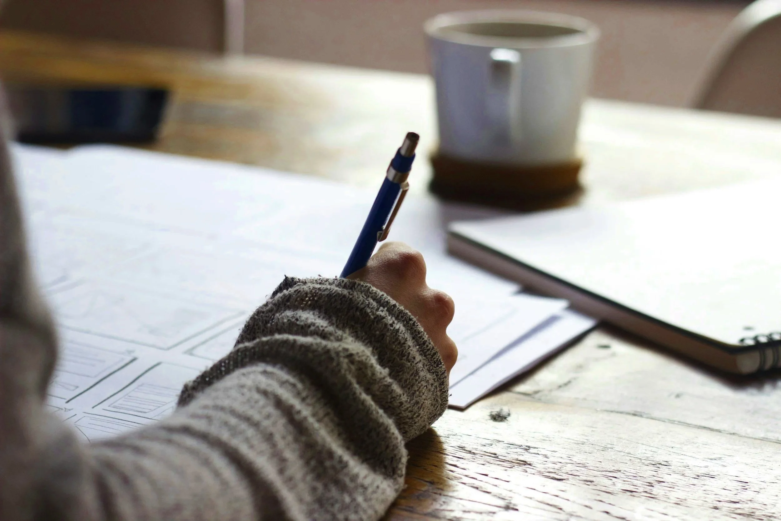 a person writing notes with a pen, a mug is sitting across the table