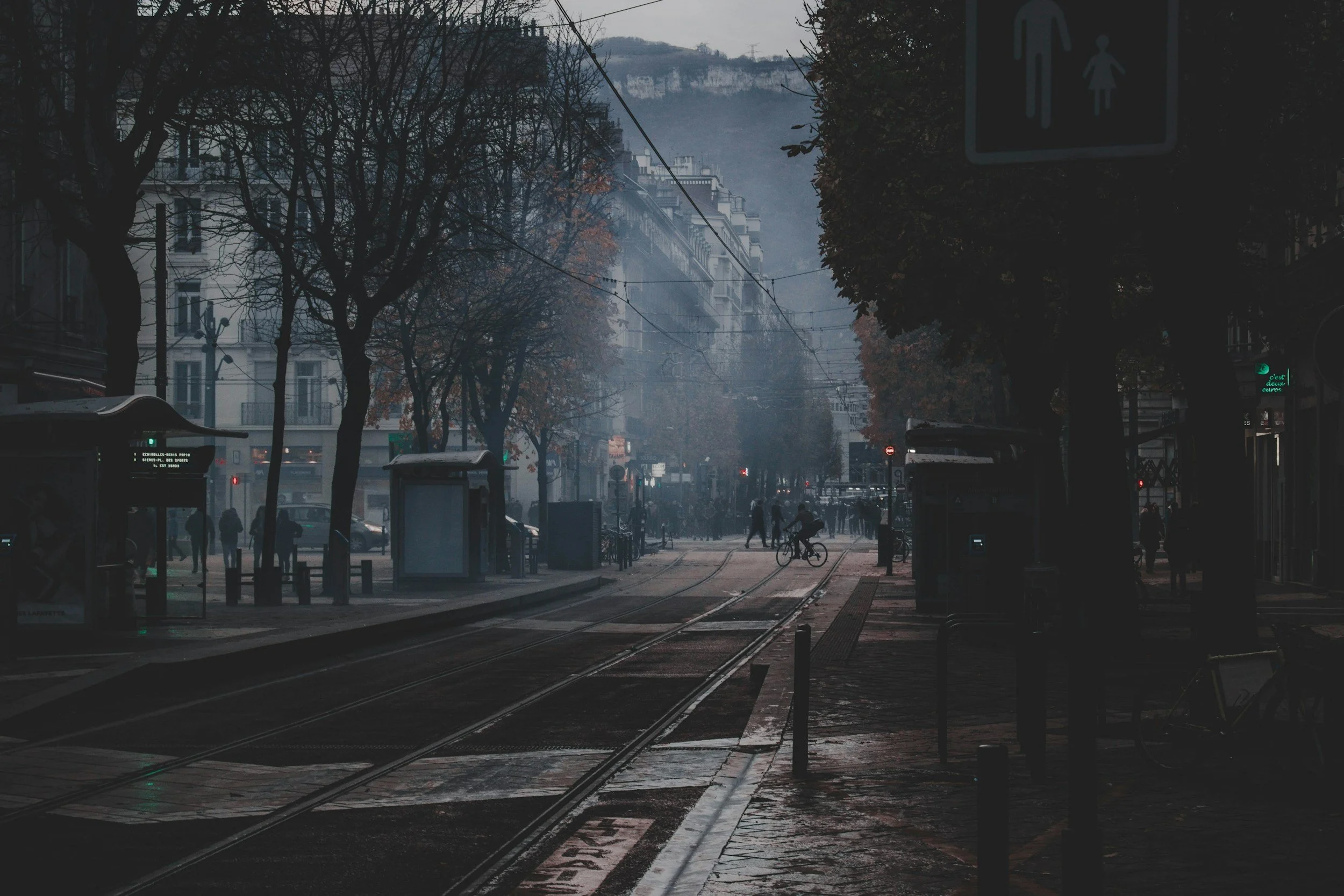 A city street scene with tram tracks, trees on both sides, and pedestrians, some cycling, on a misty morning or evening.