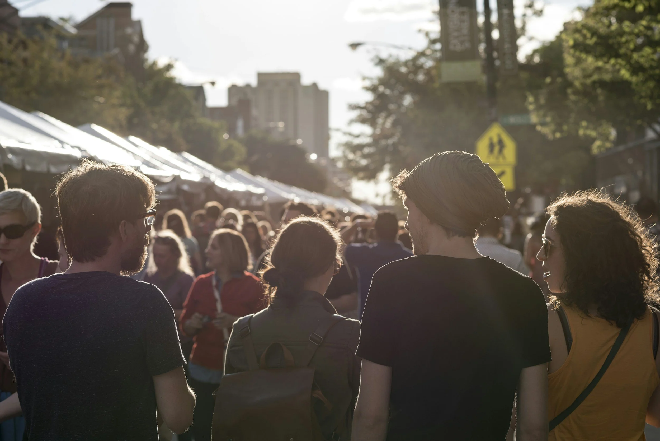 People walking and socializing at an outdoor event or festival on a sunny day with tents and city buildings in the background.