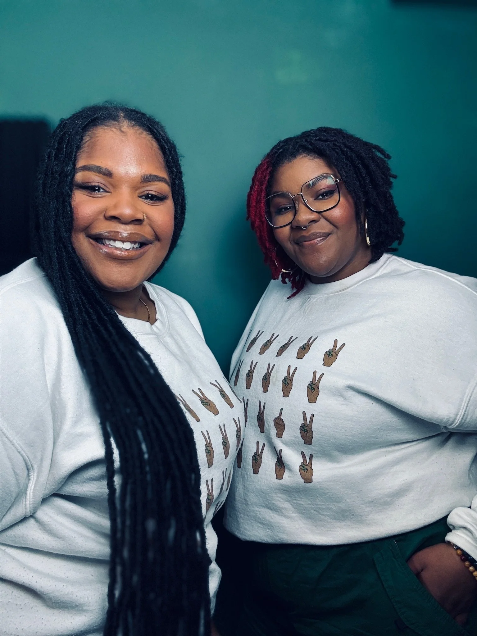 Two women standing close together, both wearing white sweatshirts with peace hand emojis, smiling at the camera against a teal background.