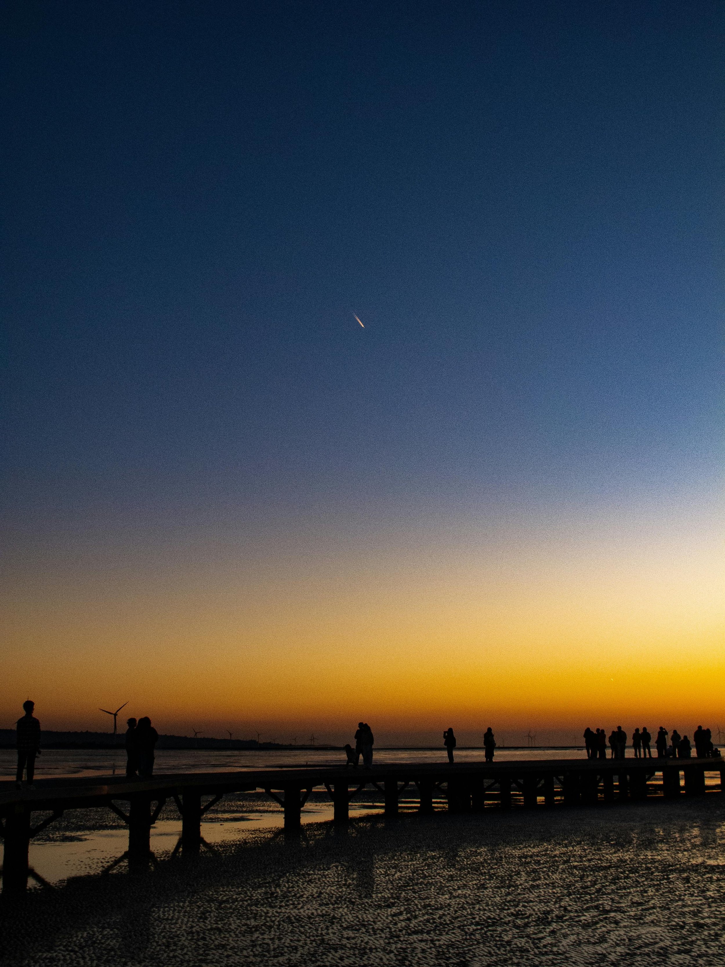 Silhouettes of people standing on a pier at sunset by the water, with wind turbines in the distance and a jet streaking across the sky.
