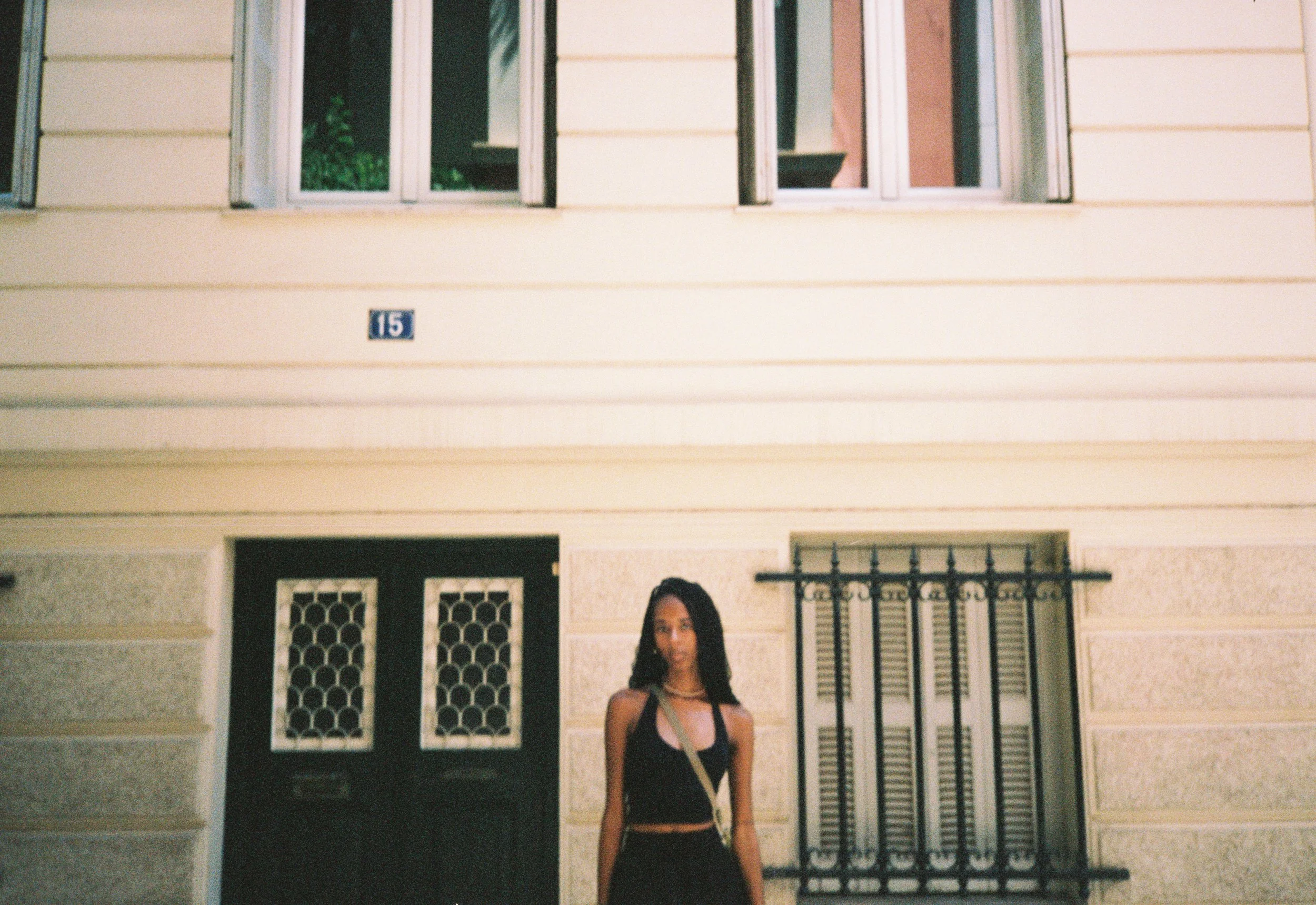 A young woman with long dark hair standing in front of a beige house with a black front door and window shutters. She is wearing a black sleeveless top and a tote bag.