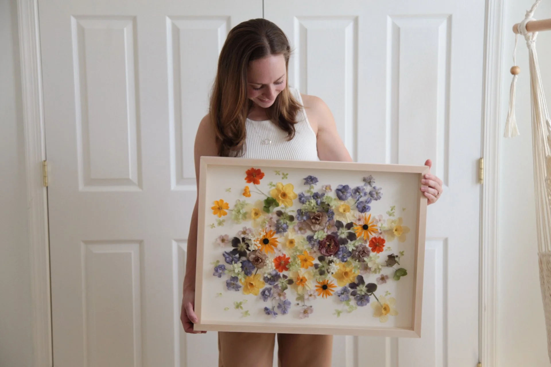 A woman with brown hair in a white sleeveless top holding a framed collage of various colorful pressed flowers, standing in front of a white door with paneled design.