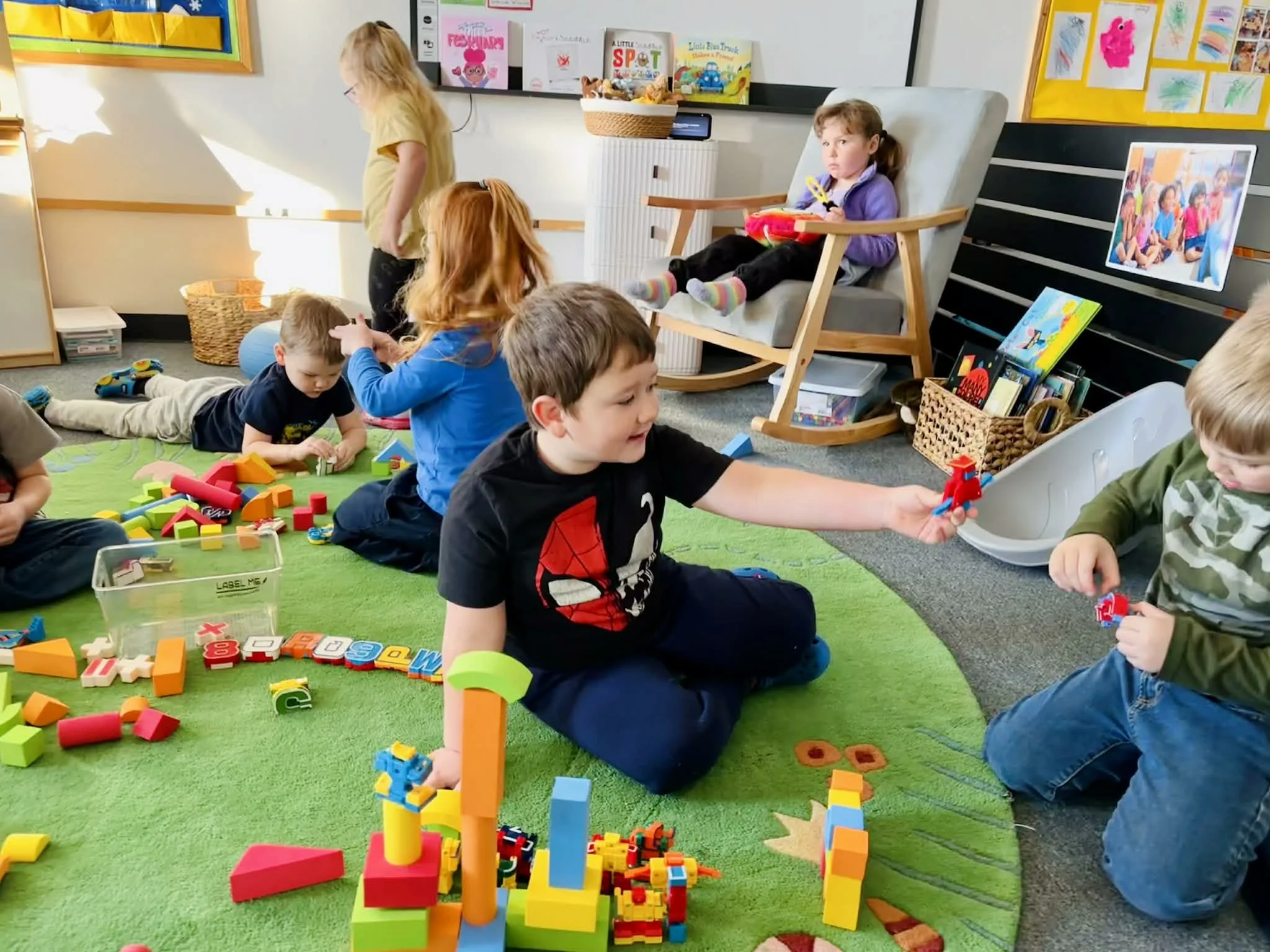 Children Playing on a green circle rug in a classroom with colorful blocks and sharing together.