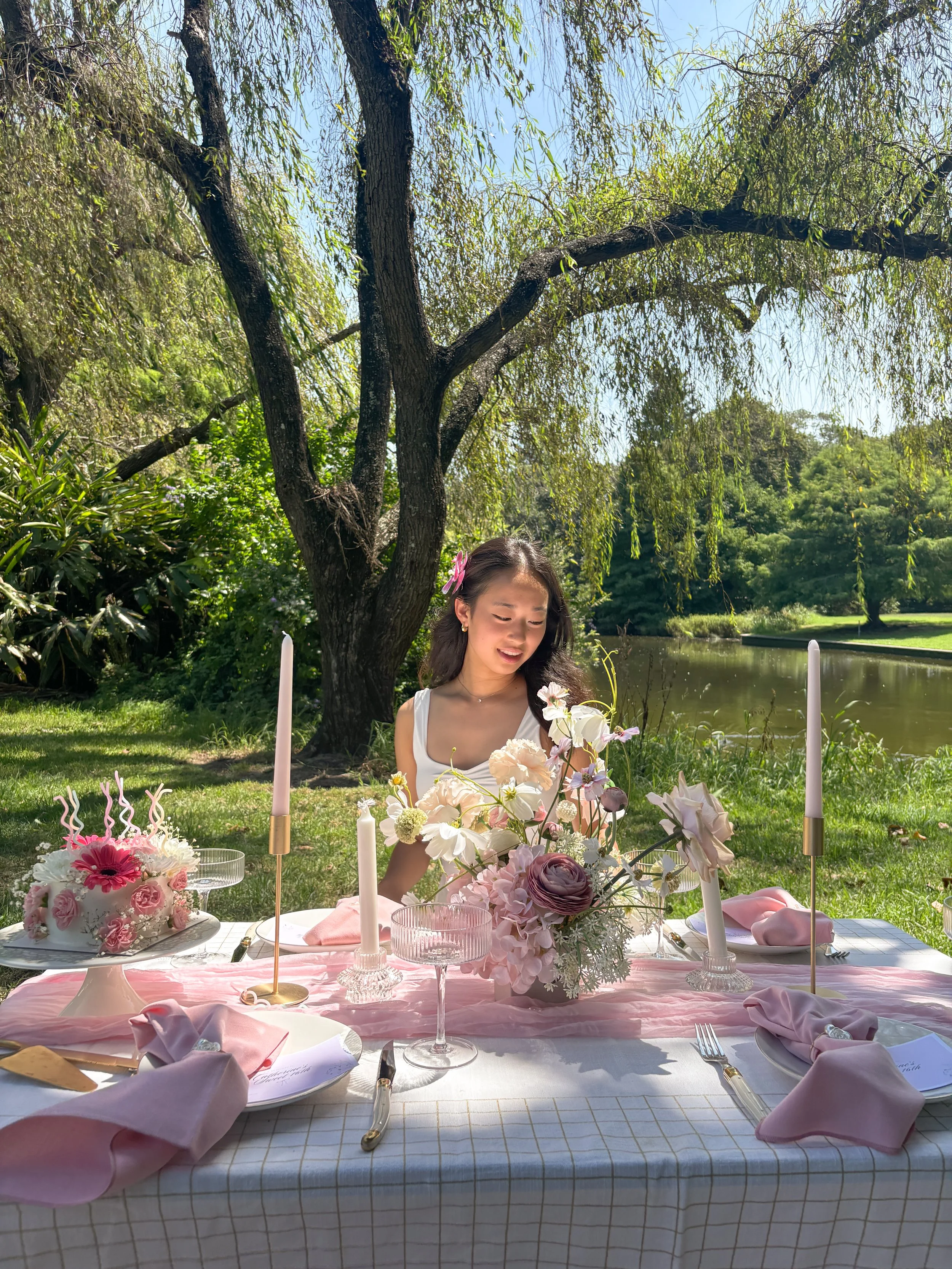A young woman sitting at an outdoor table in a lush green park near a river, decorated with pink and white flowers, pink napkins, candles, and elegant tableware, enjoying a sunny day.
