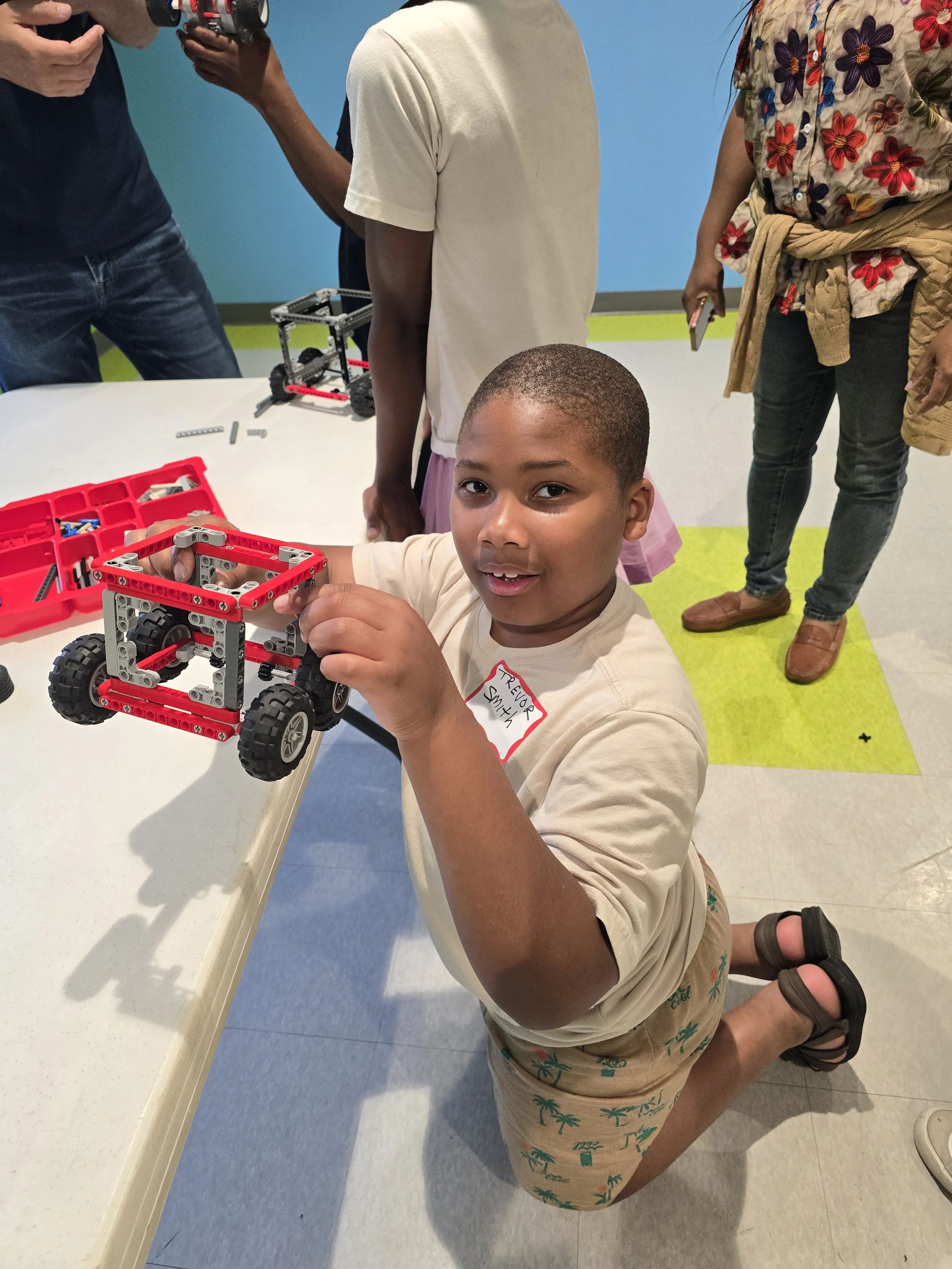 A young boy kneels on the floor, playing with a red and gray LEGO vehicle at a table. Other children and adults stand around, observing or holding devices. The setting appears to be an indoor activity or educational space with colorful walls and flooring.
