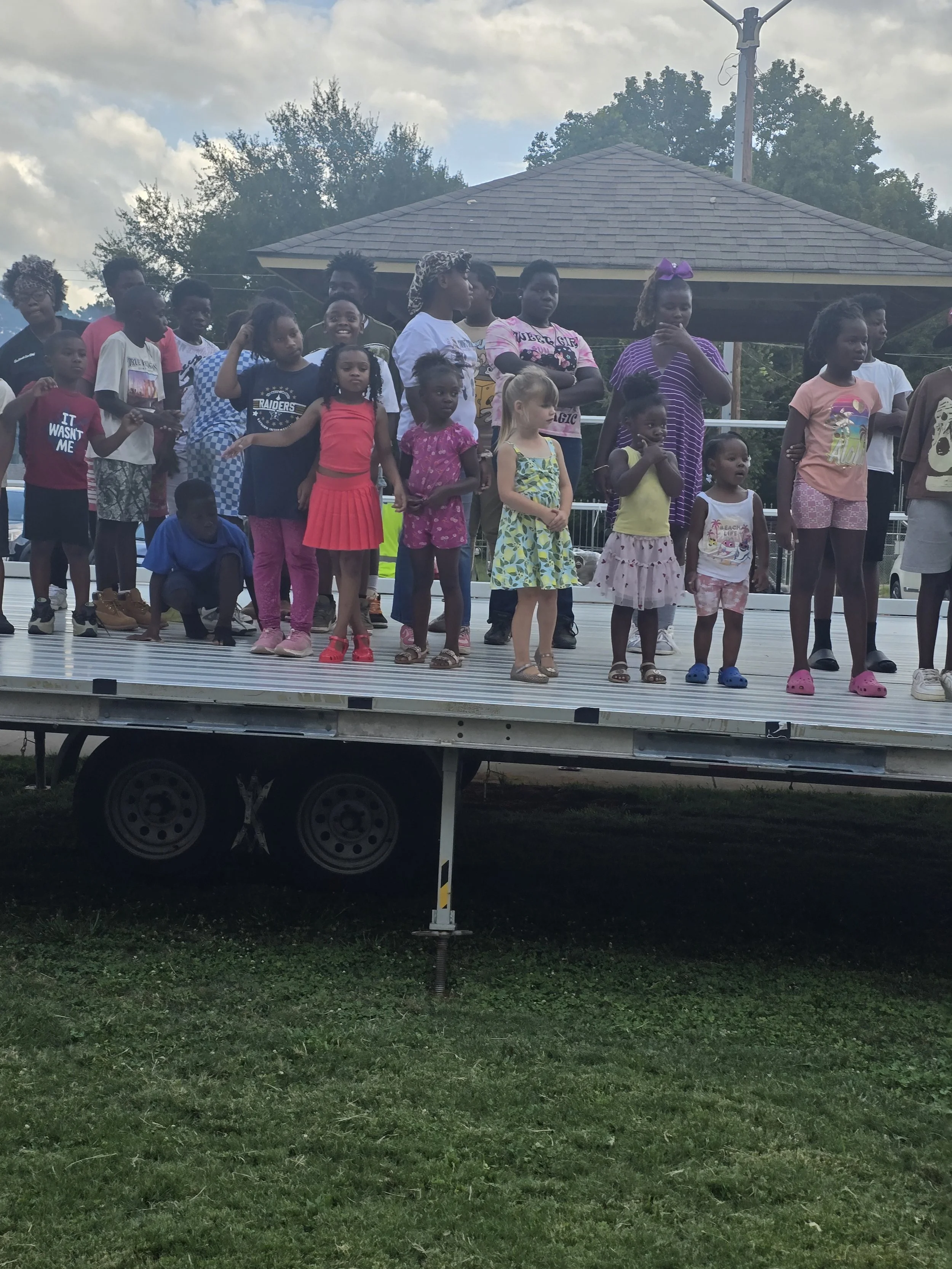 Group of children gathered on a stage outdoors, some standing and some sitting, with a pavilion, trees, and cloudy sky in the background.
