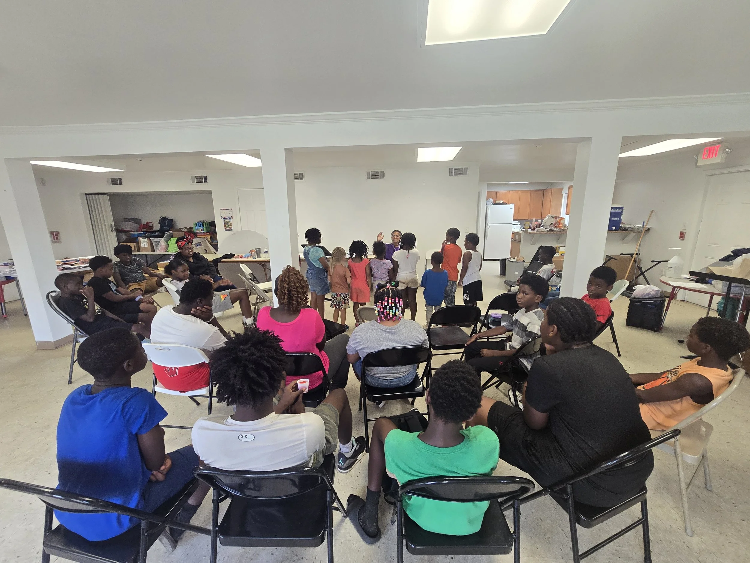 A group of children and a few adults gathered in a room with white walls, some sitting on chairs and others standing, engaging in an activity or discussion.