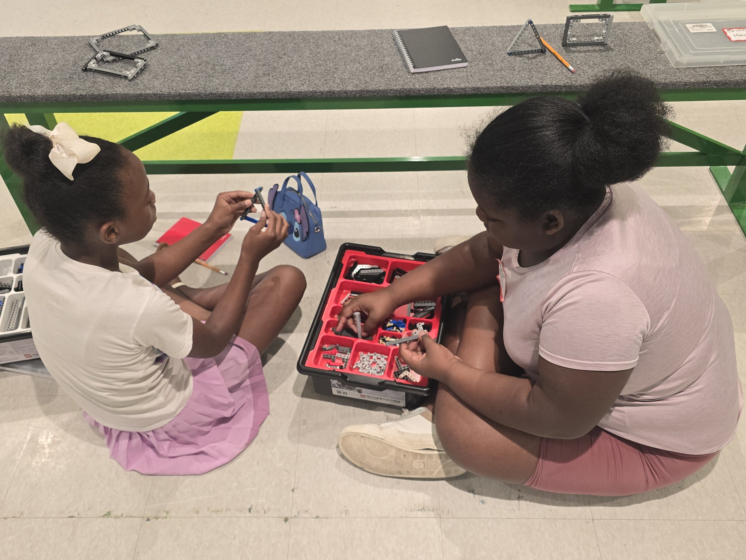 A young girl and woman sitting on the floor assembling LEGO robotics. The girl holds a LEGO piece while the woman reaches into a red container of LEGO parts. There is a green table with a gray felt top and LEGO structures on it.