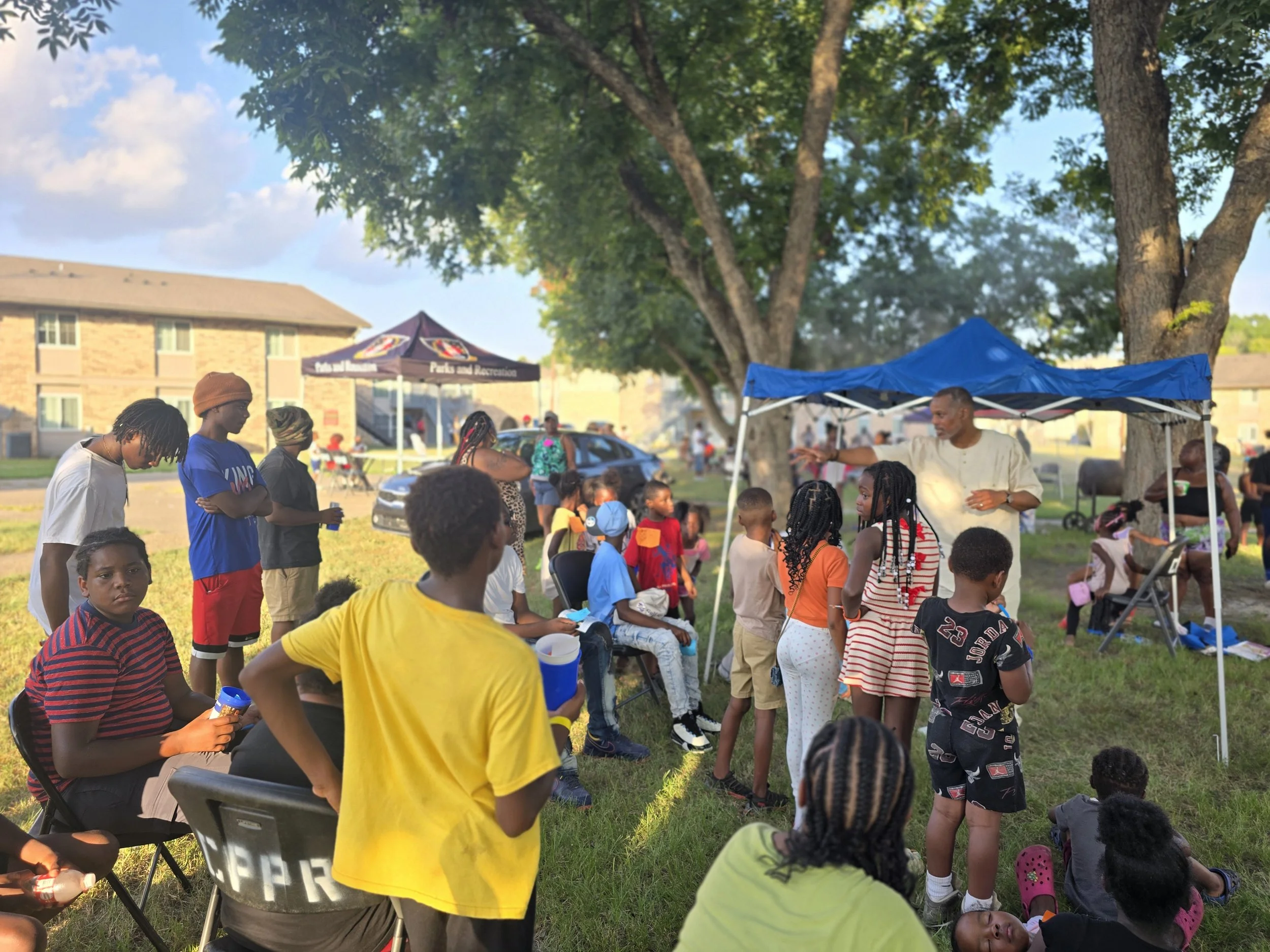 A large gathering of children and adults outdoors under trees, with some seated and others standing, near tents and a building, sunlight filtering through the leaves.