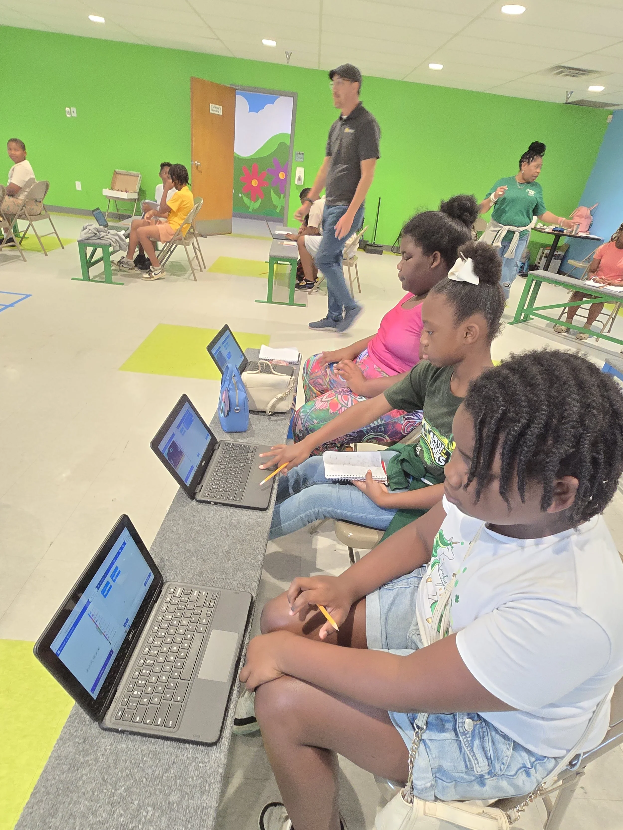 Children sitting on chairs in a classroom with bright green walls, some children working on laptops, and a teacher standing near a table.