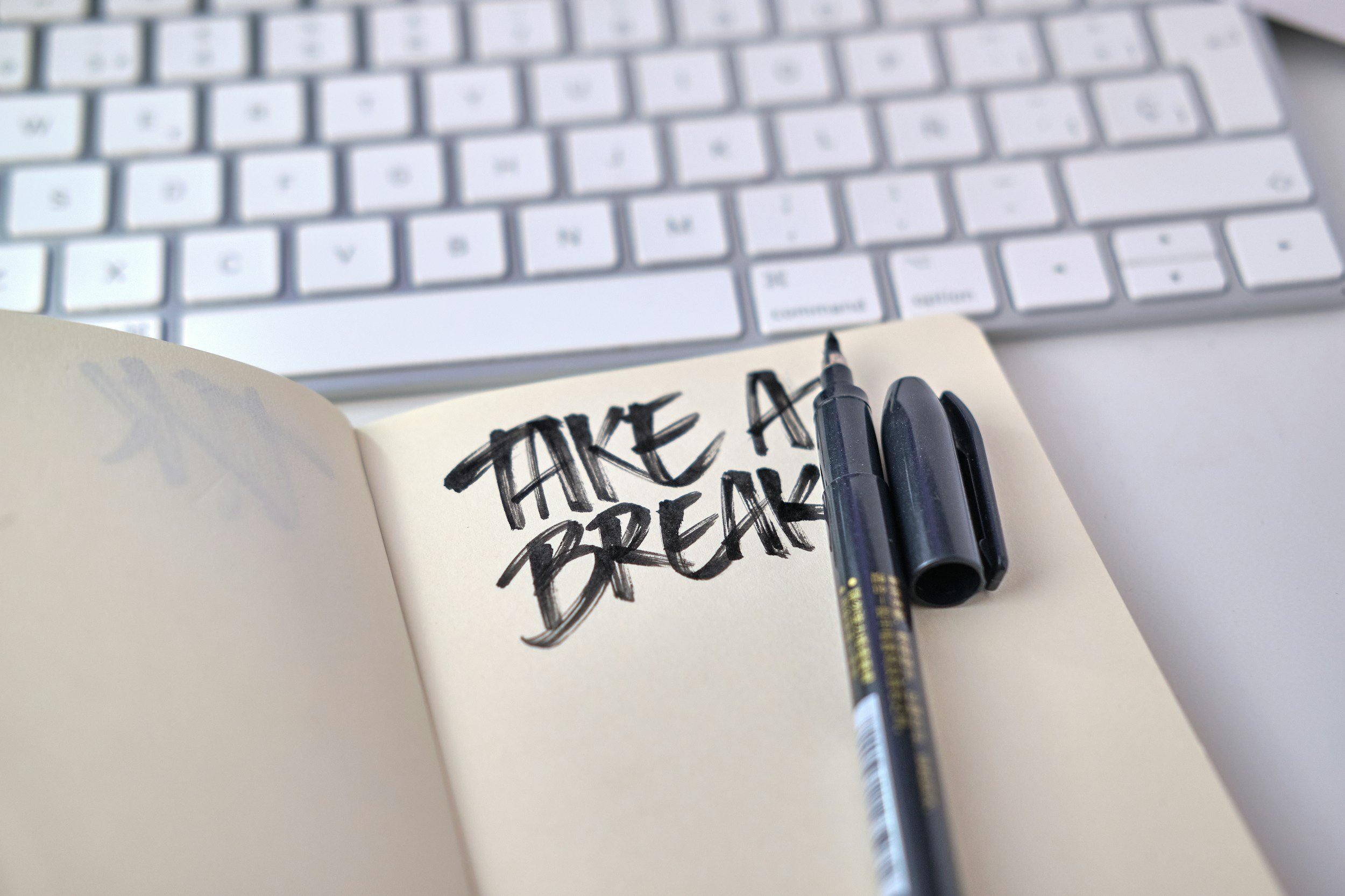 A notebook with the words 'Take a Break' written in bold black marker, placed on a white surface with a black marker pen beside it. In the background, there is a white computer keyboard.