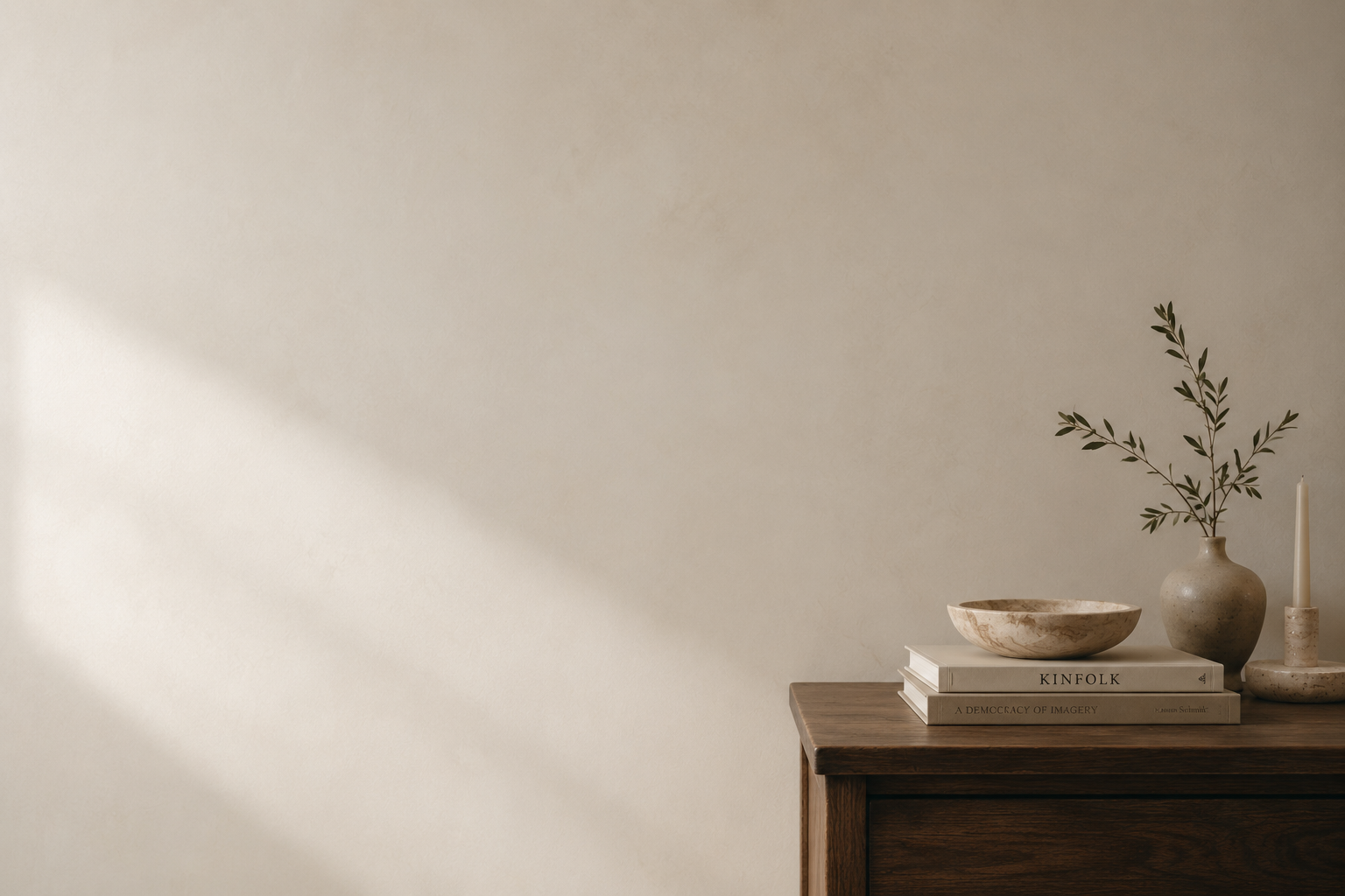 A wooden side table with a white vase holding greenery, a white candle, a stone bowl, and a couple of books on a beige wall background.